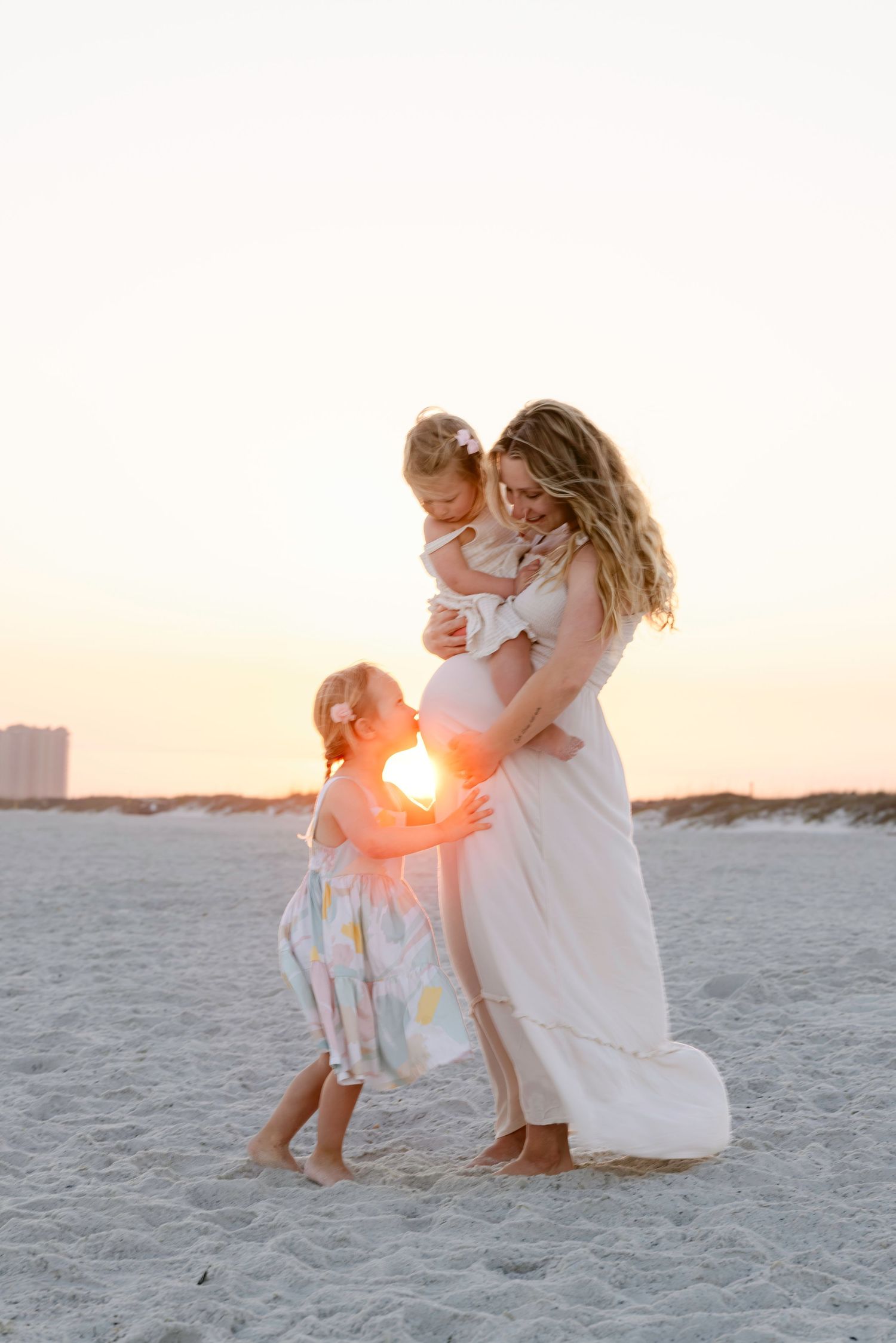 Pregnant woman in white dress holds toddler while another child hugs her belly on a sandy beach at sunset.