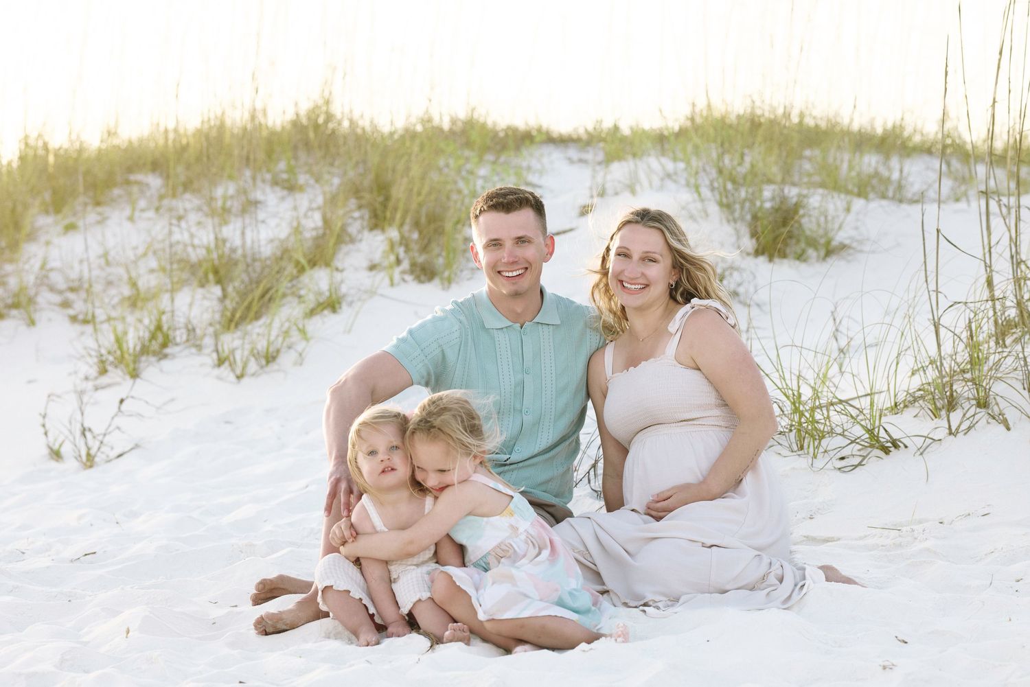 Family of four with pregnant mother and two young daughters sit together on white sand beach dunes.