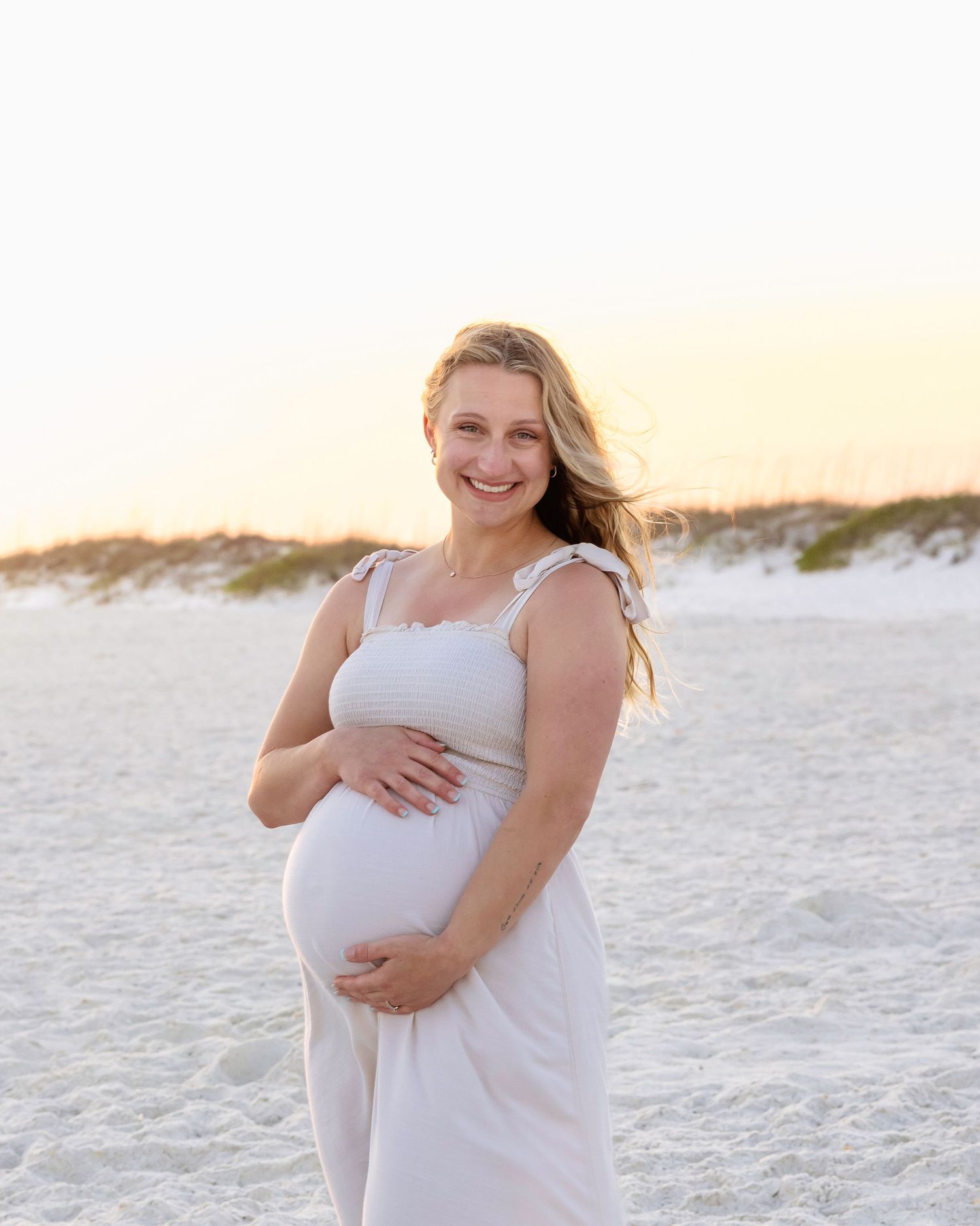 Pregnant woman in white dress cradling baby bump on white sand beach at golden hour sunset.