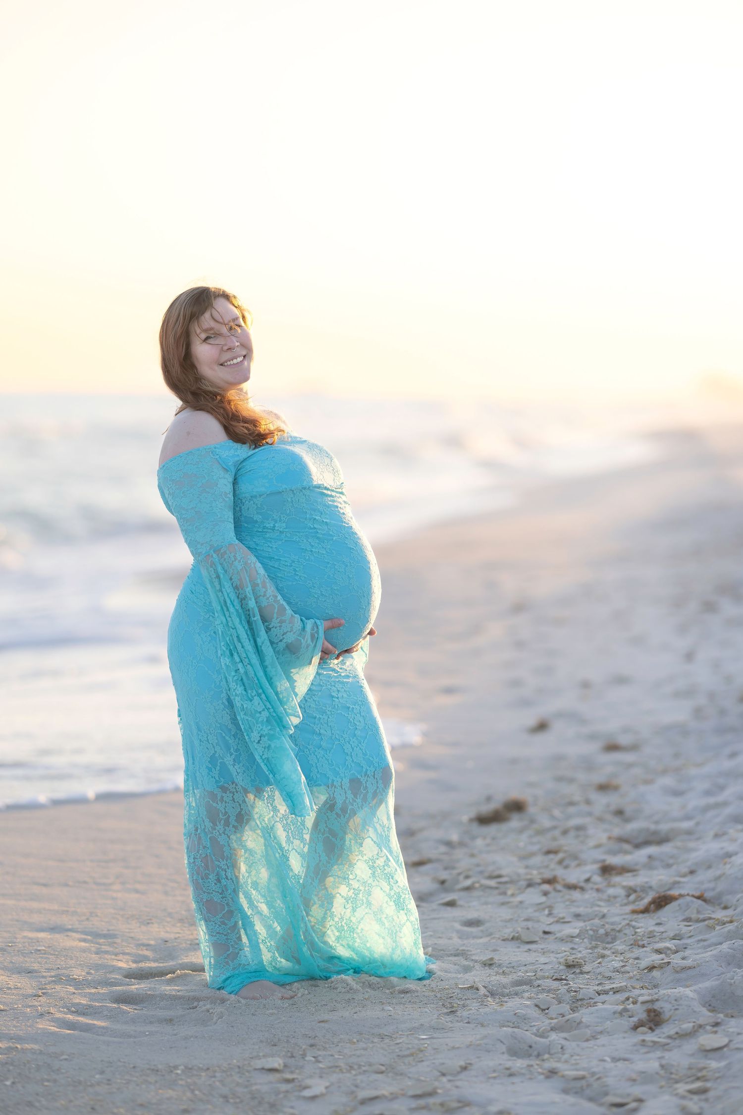 Smiling pregnant woman in teal lace gown cradling her baby bump on a sunlit beach at sunset.