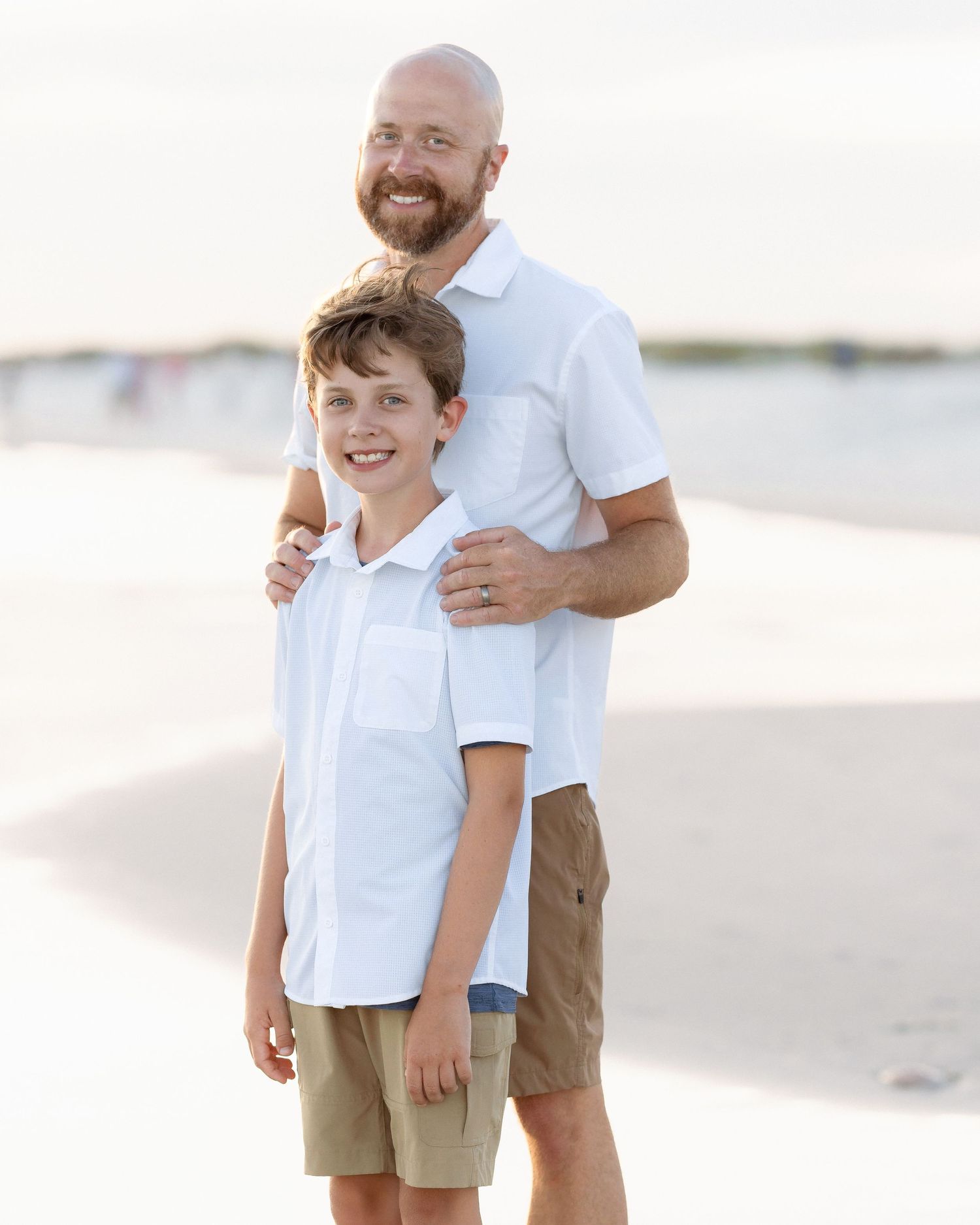 A smiling bald man with a beard stands behind a young boy on a sandy beach, both wearing white shirts.