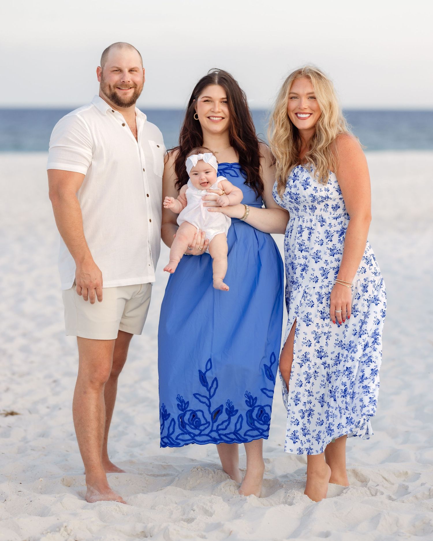 Family of three adults and a baby posing on a sunny beach, wearing blue and white outfits.