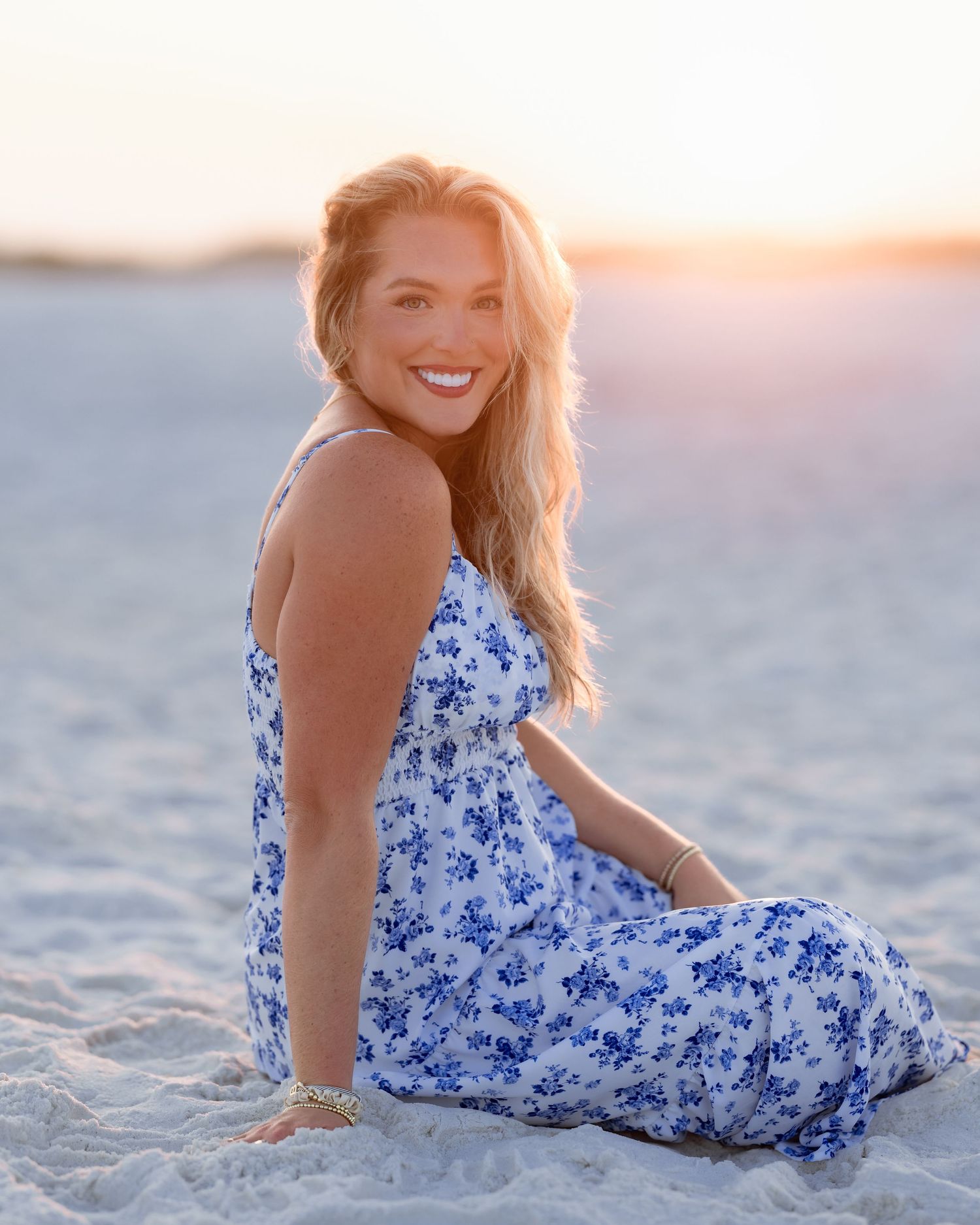 Smiling woman in blue floral sundress sitting on sandy beach at golden sunset hour.