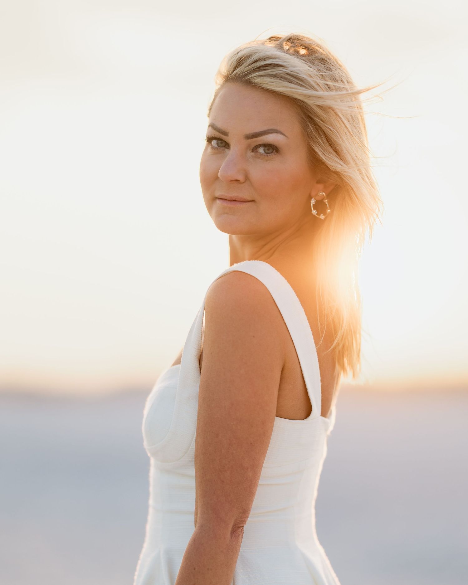 Blonde woman in white dress with gold hoop earrings posing at golden hour on beach.