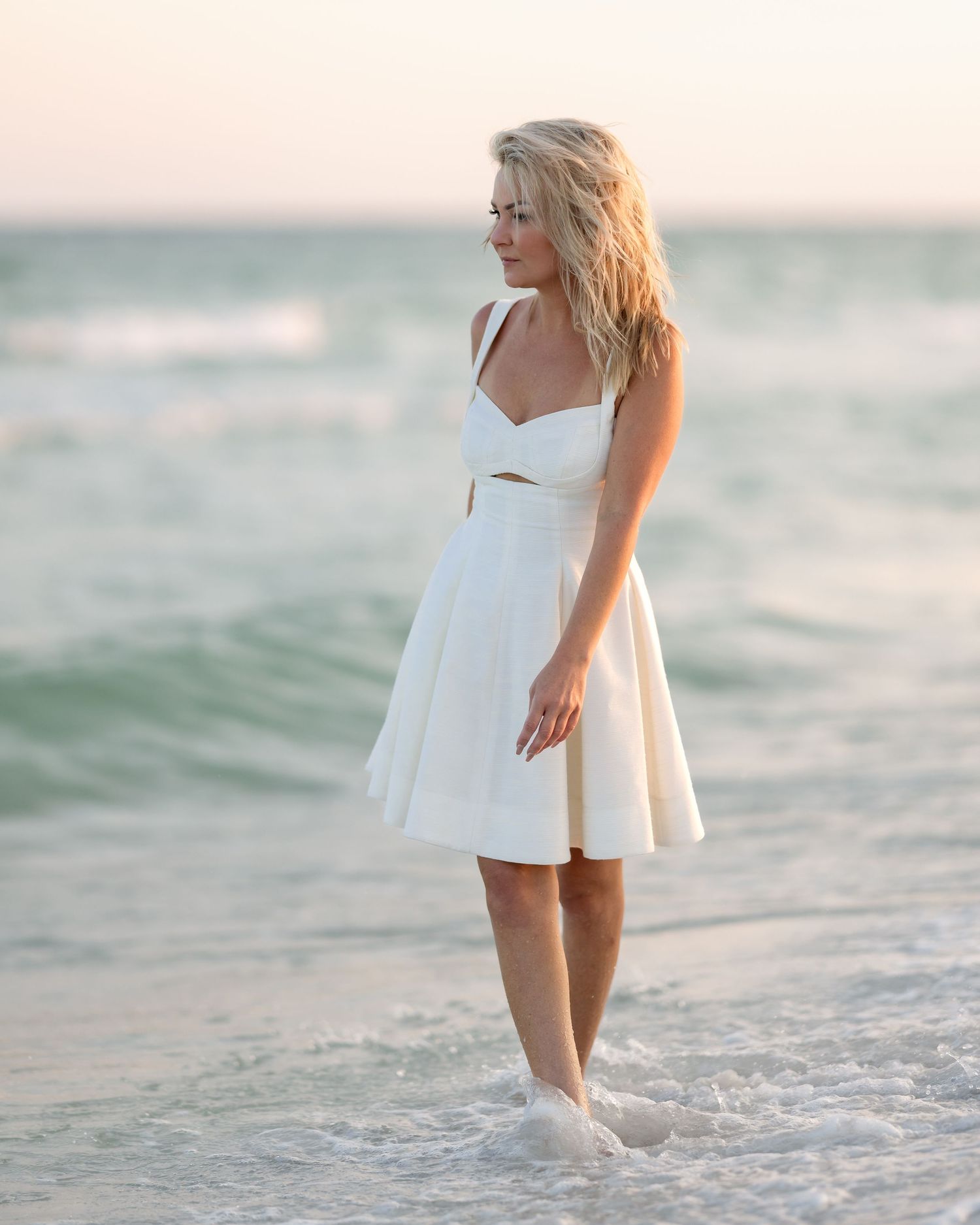 Blonde woman in white sundress standing in shallow ocean waves at sunset beach.
