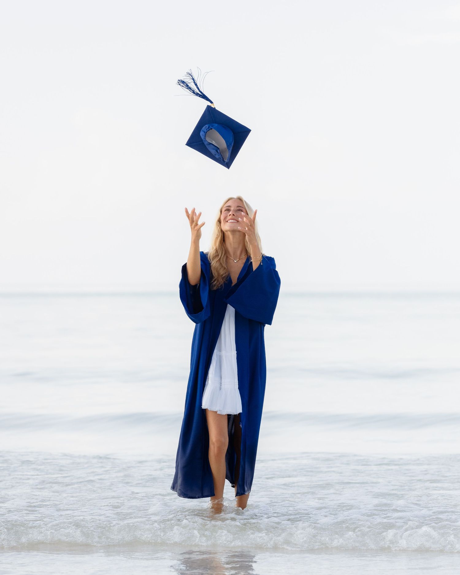 A joyful graduate in a blue gown tosses her cap on a beach, celebrating her achievement by the ocean.