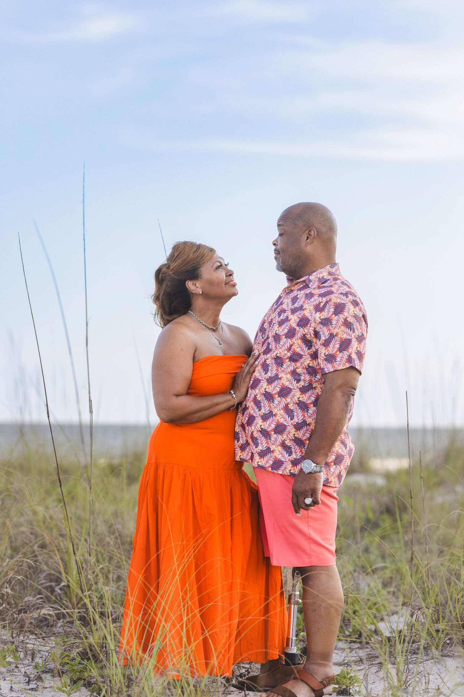 Couple in colorful outfits sharing a romantic moment on a sandy beach with sea grass and blue sky.