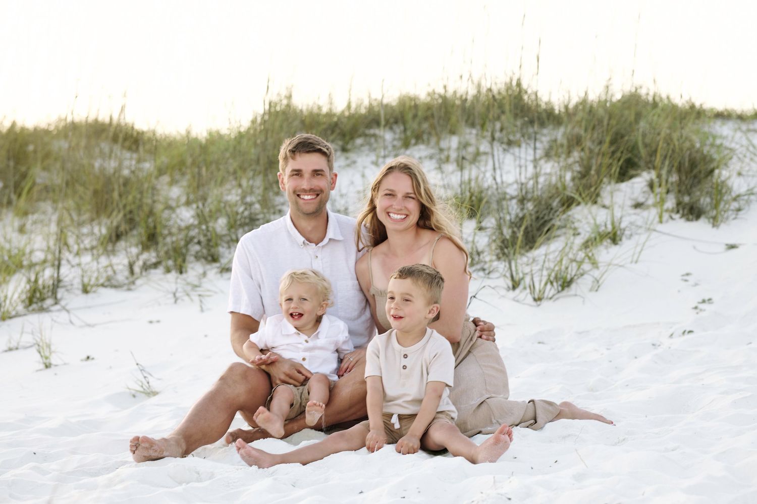 Family of four smiling together on white sand beach dunes, parents with two young boys in coordinating neutral outfits.