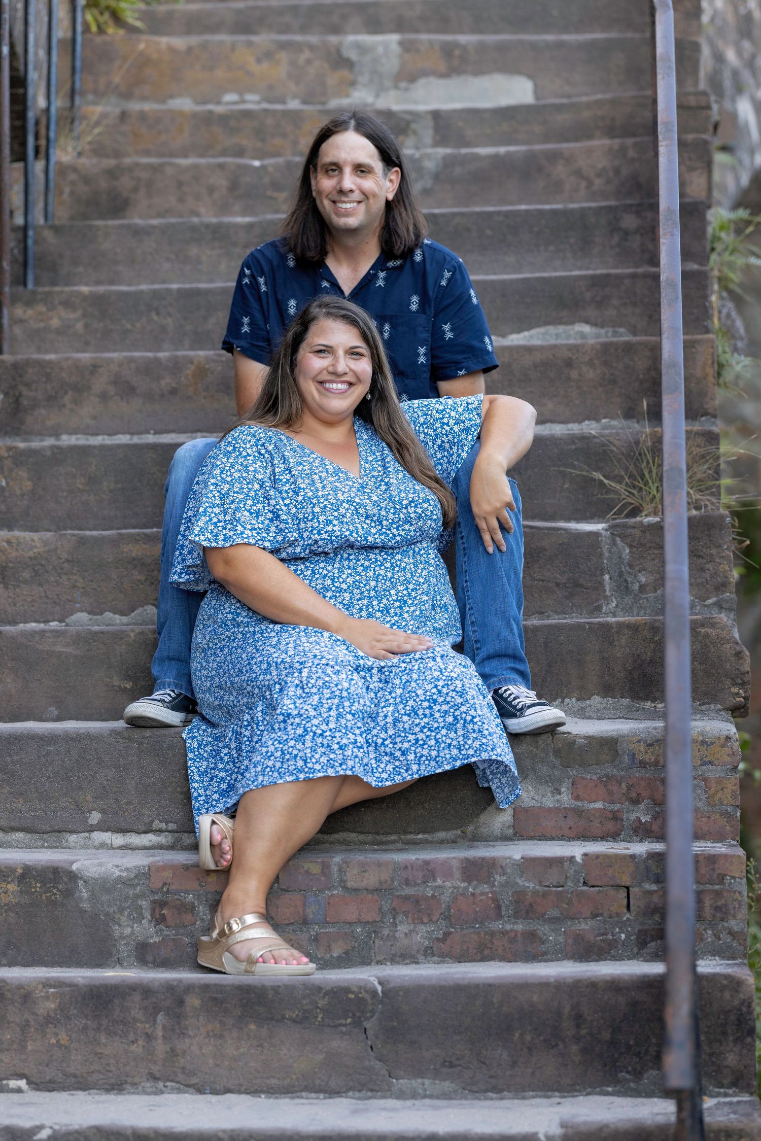 Smiling pregnant woman in blue floral dress sits on stone steps with long-haired man in blue shirt behind her.