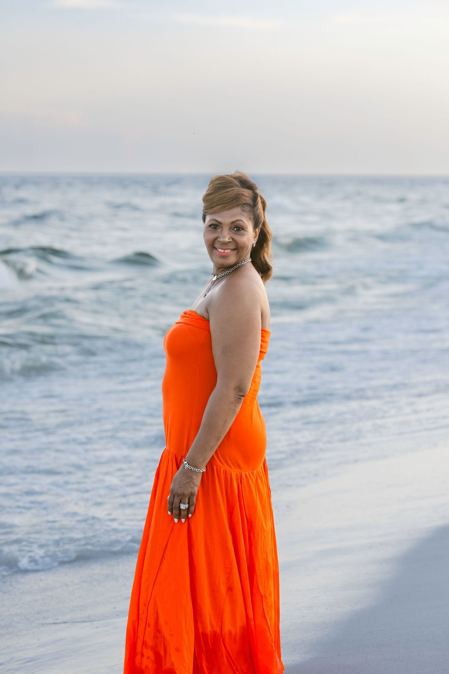 Smiling woman in strapless orange maxi dress poses on beach with ocean waves behind her.