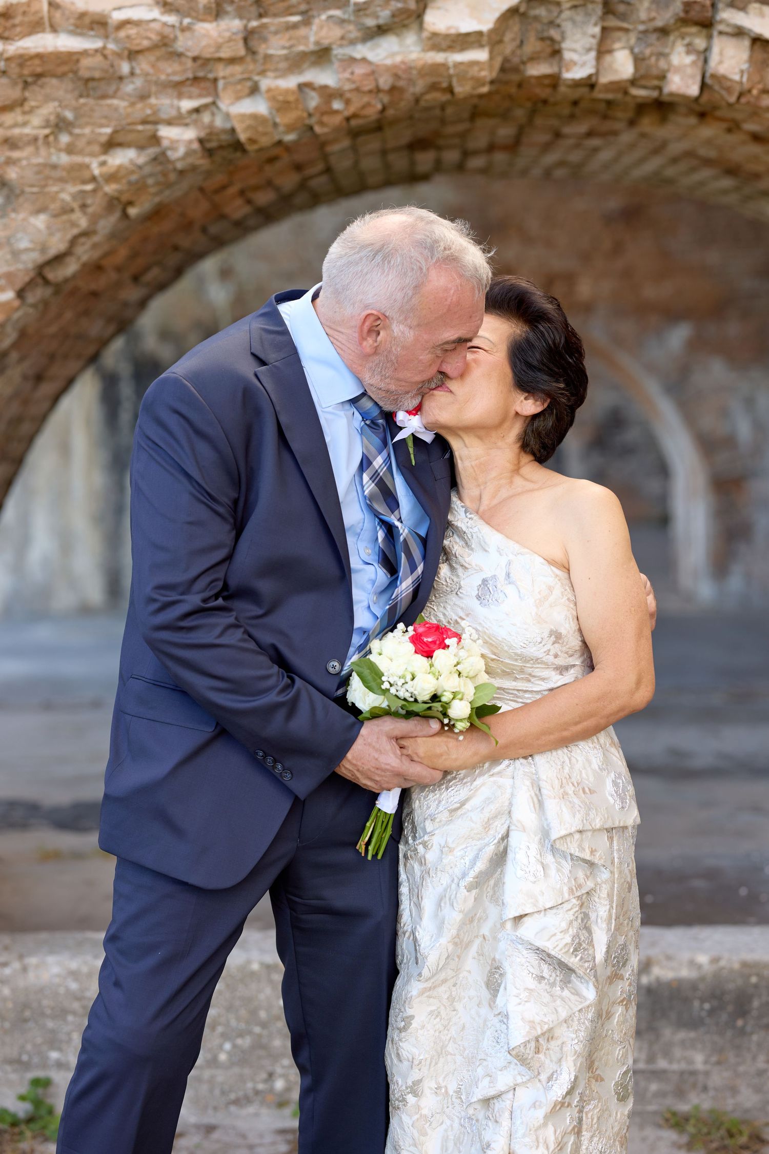 Mature interracial couple sharing a kiss on their wedding day under a stone arch.