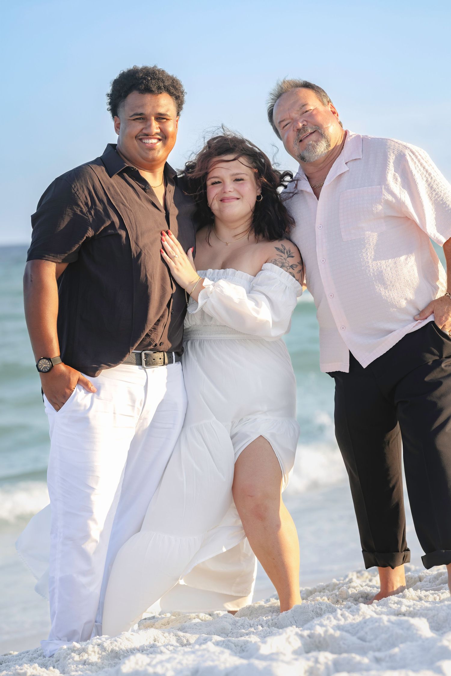 Three people smiling on a sunny beach, woman in white dress flanked by two men in casual attire.