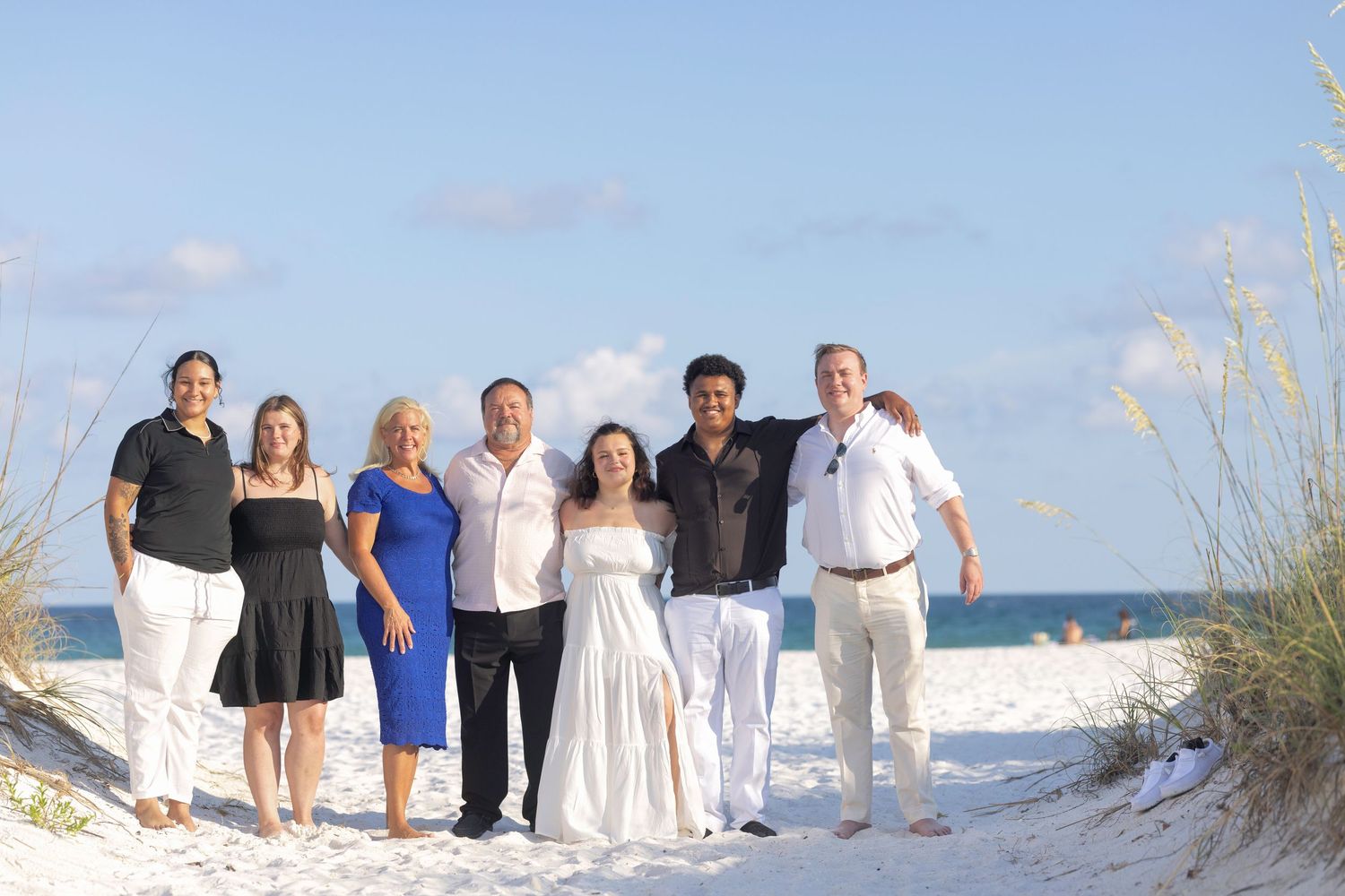 Group of seven people smiling together on a sunny white sand beach with sea oats and blue sky.