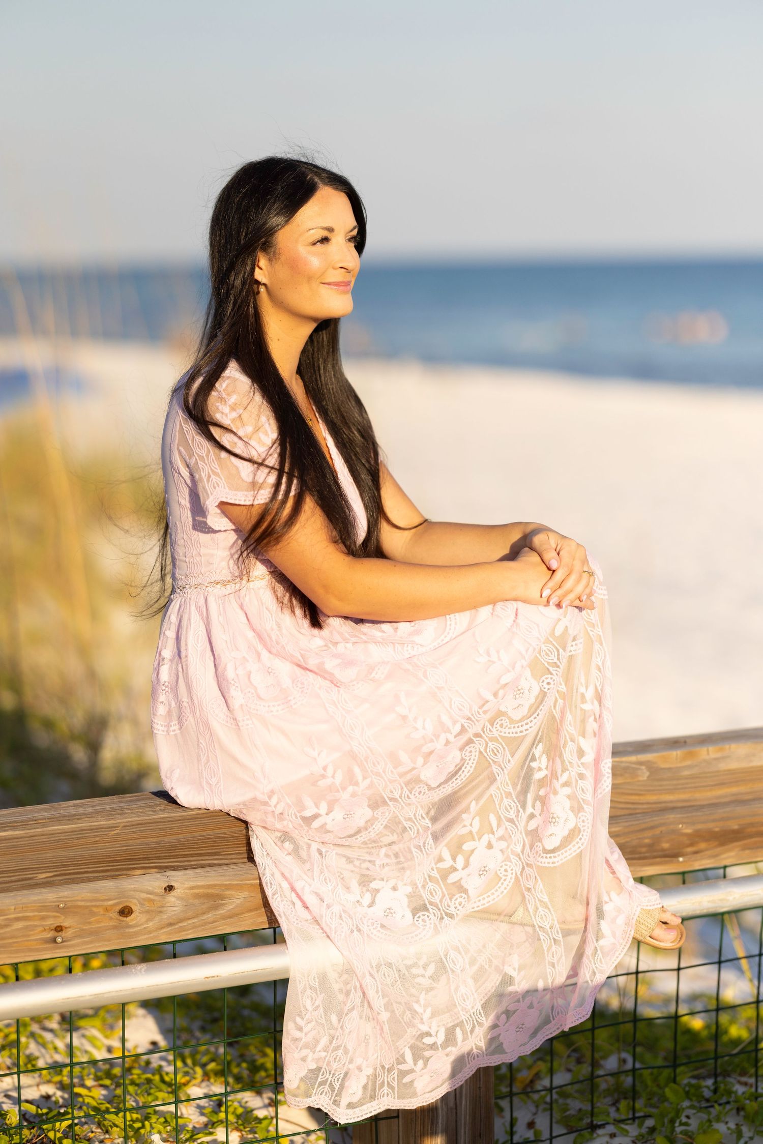 Woman in pink lace dress sitting on beach fence at sunset.