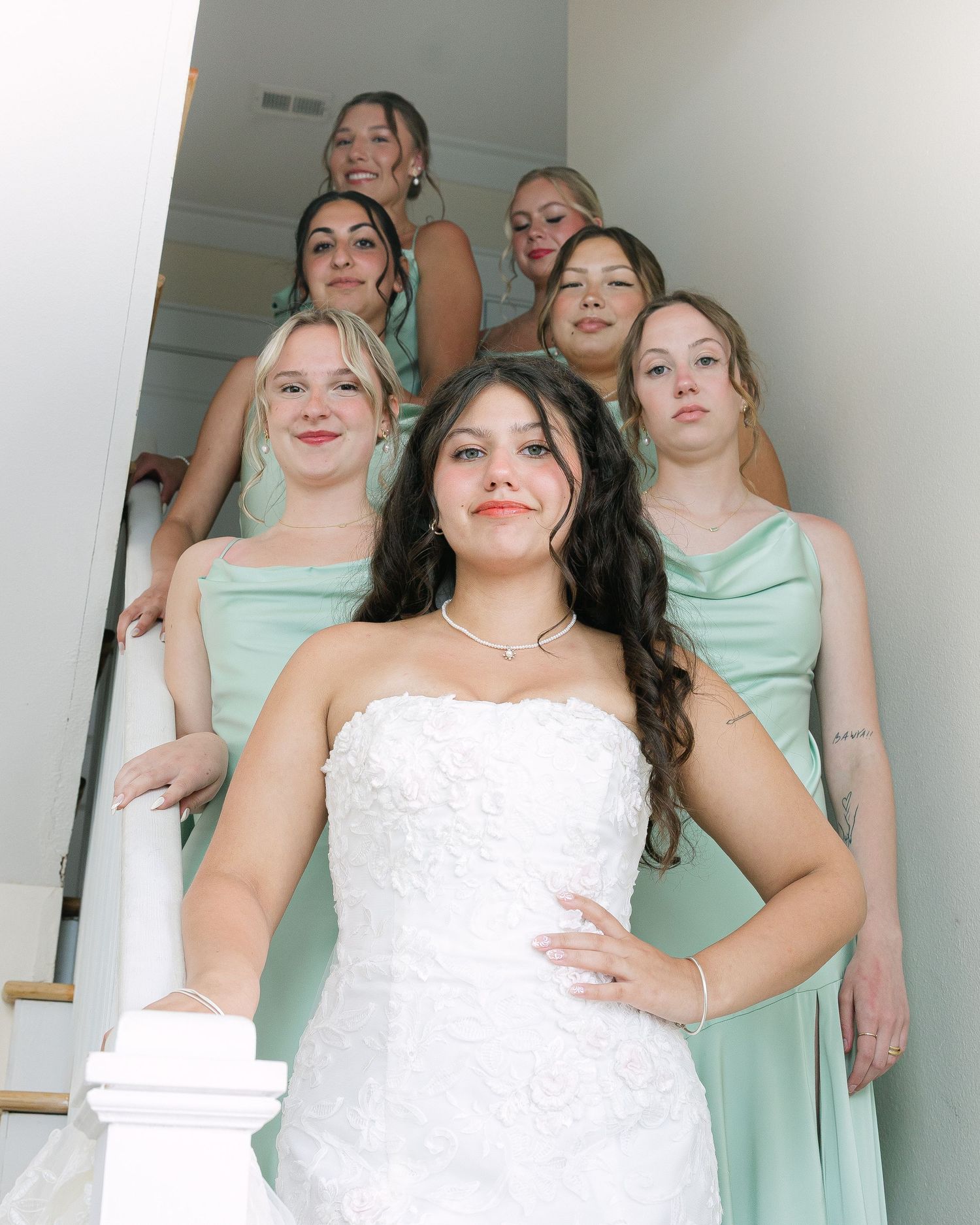 Bride in white strapless gown poses with six bridesmaids in sage green dresses on elegant staircase.