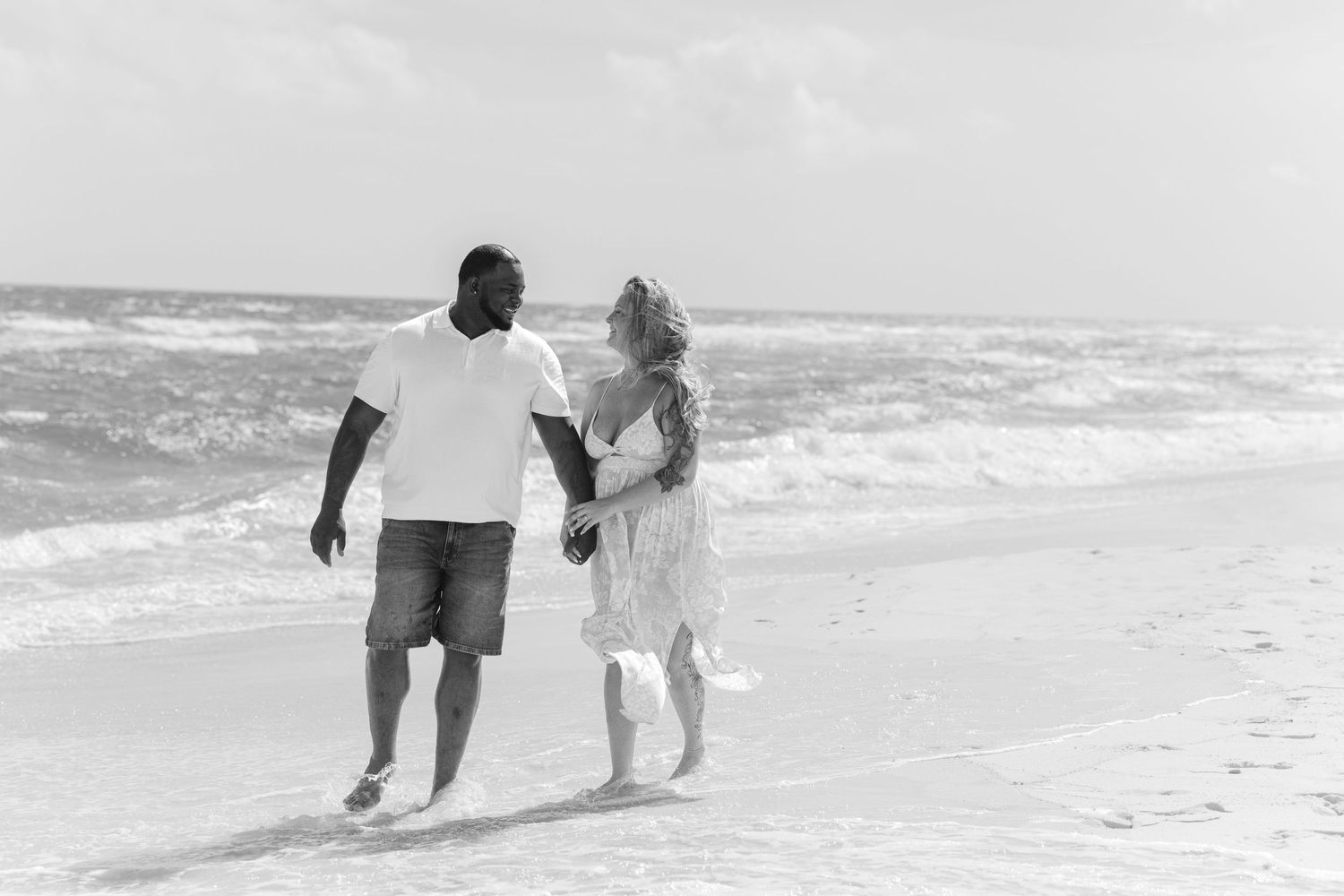 Black and white photo of interracial couple holding hands and walking along a sunny beach shoreline.