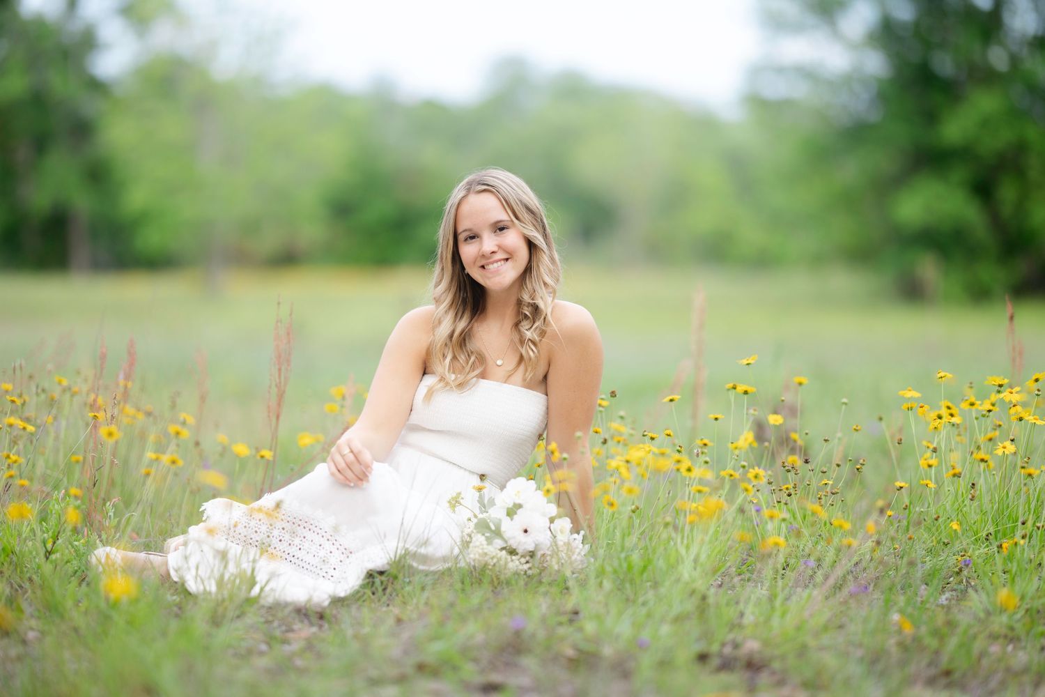 Young woman in white dress sitting in a wildflower meadow with yellow flowers, smiling outdoors.