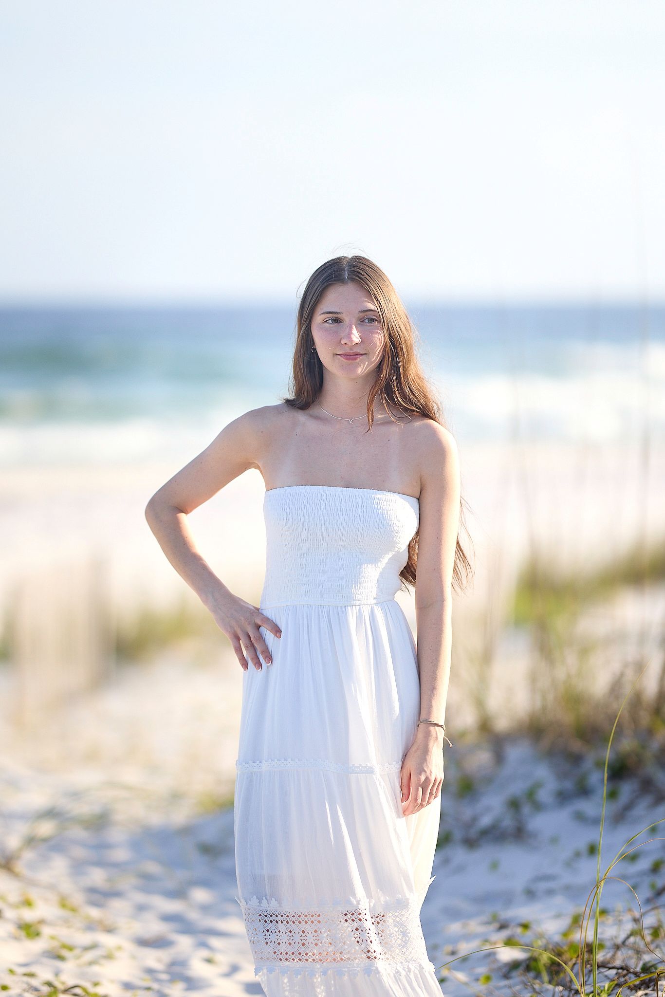 Young woman in white strapless maxi dress posing on sandy beach with ocean waves in background.
