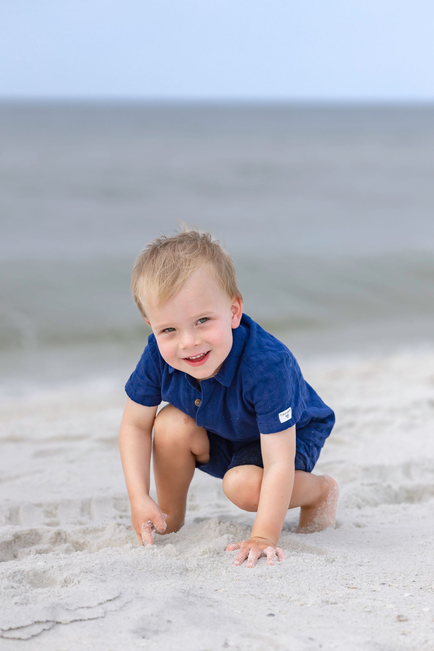 Happy toddler boy in blue shirt crouching and playing in white sand at the beach.