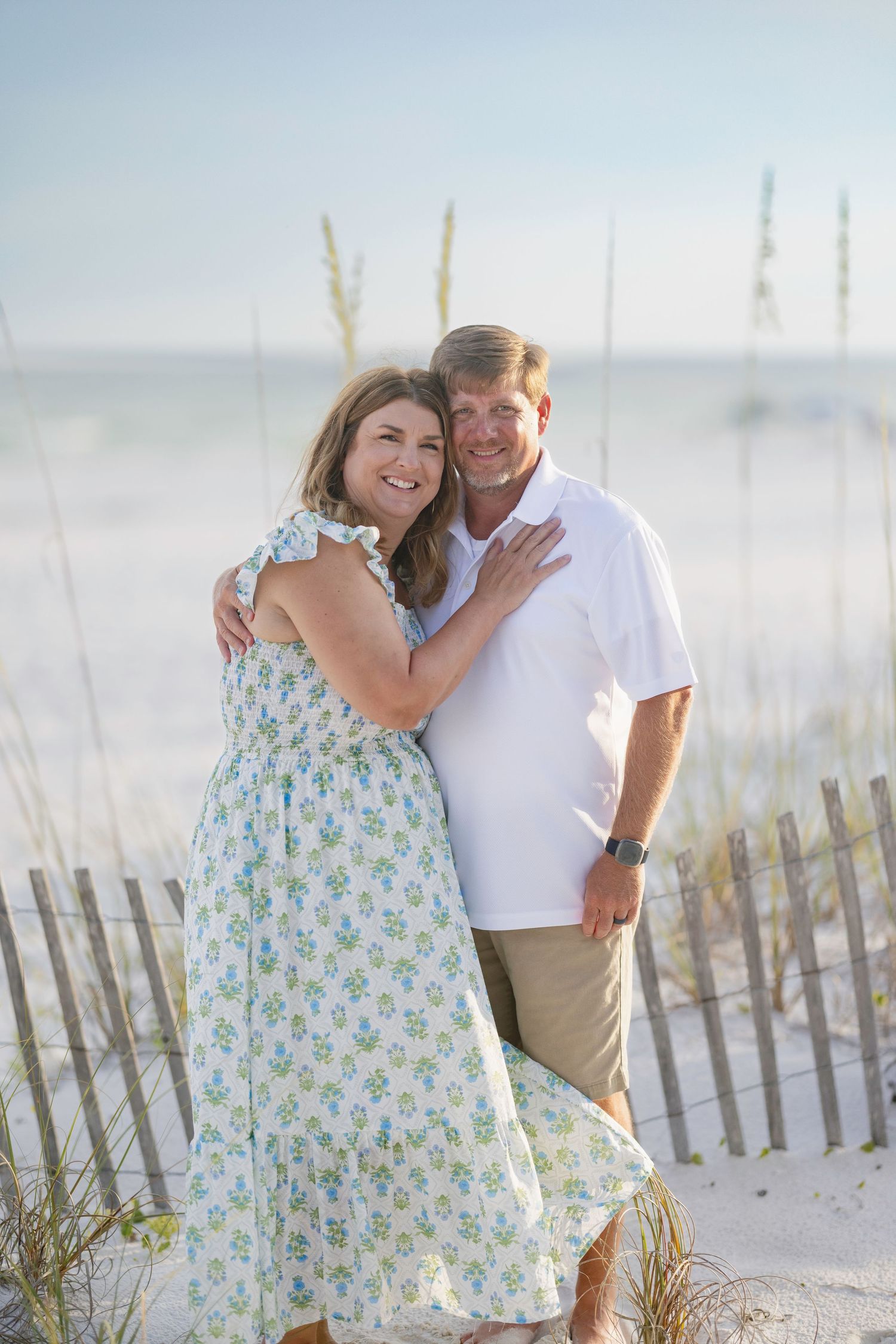 Smiling couple embracing on a sunny beach with sea oats and ocean in the background.