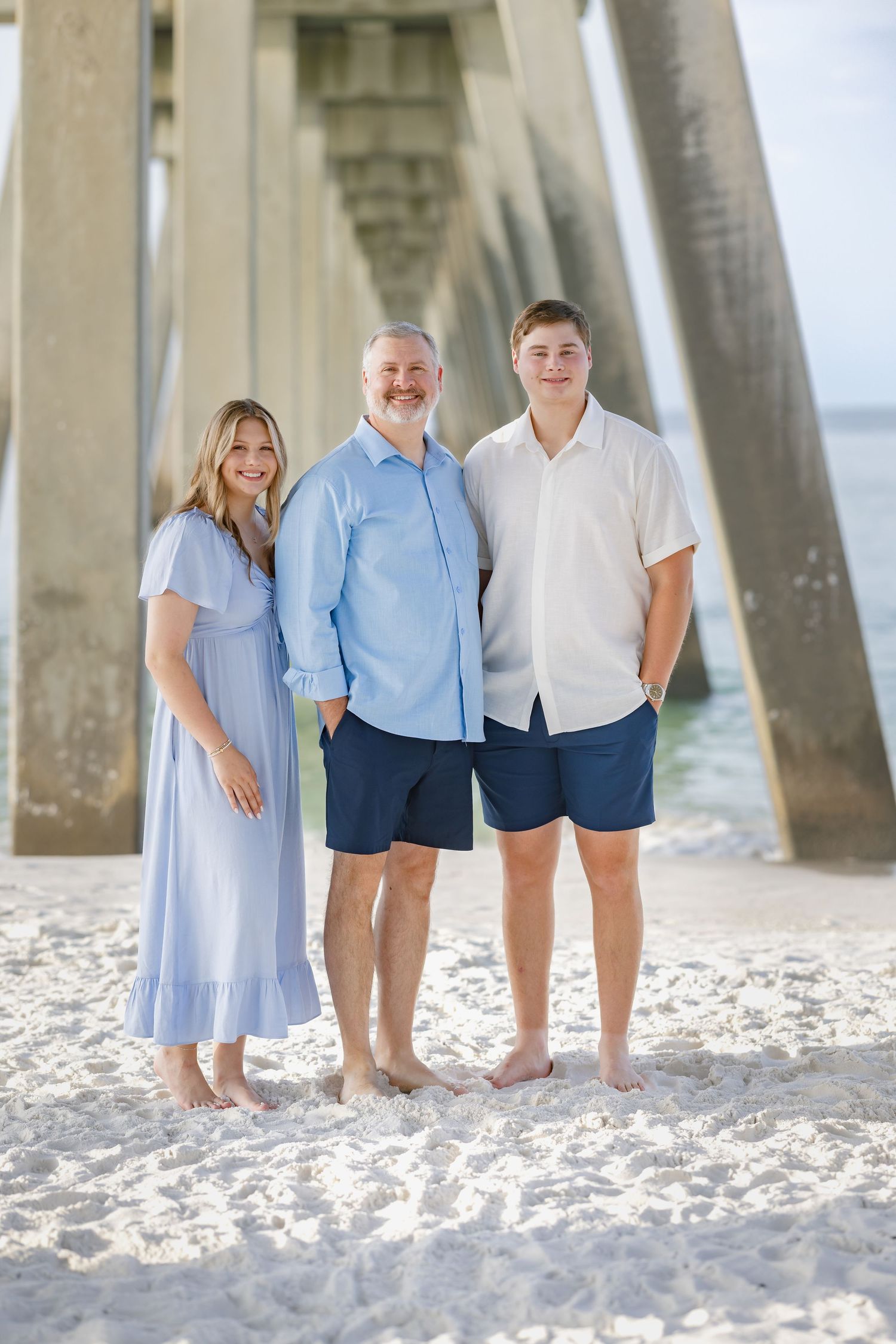 Family of three posing barefoot on white sand beach under pier, wearing coordinated blue and white outfits.