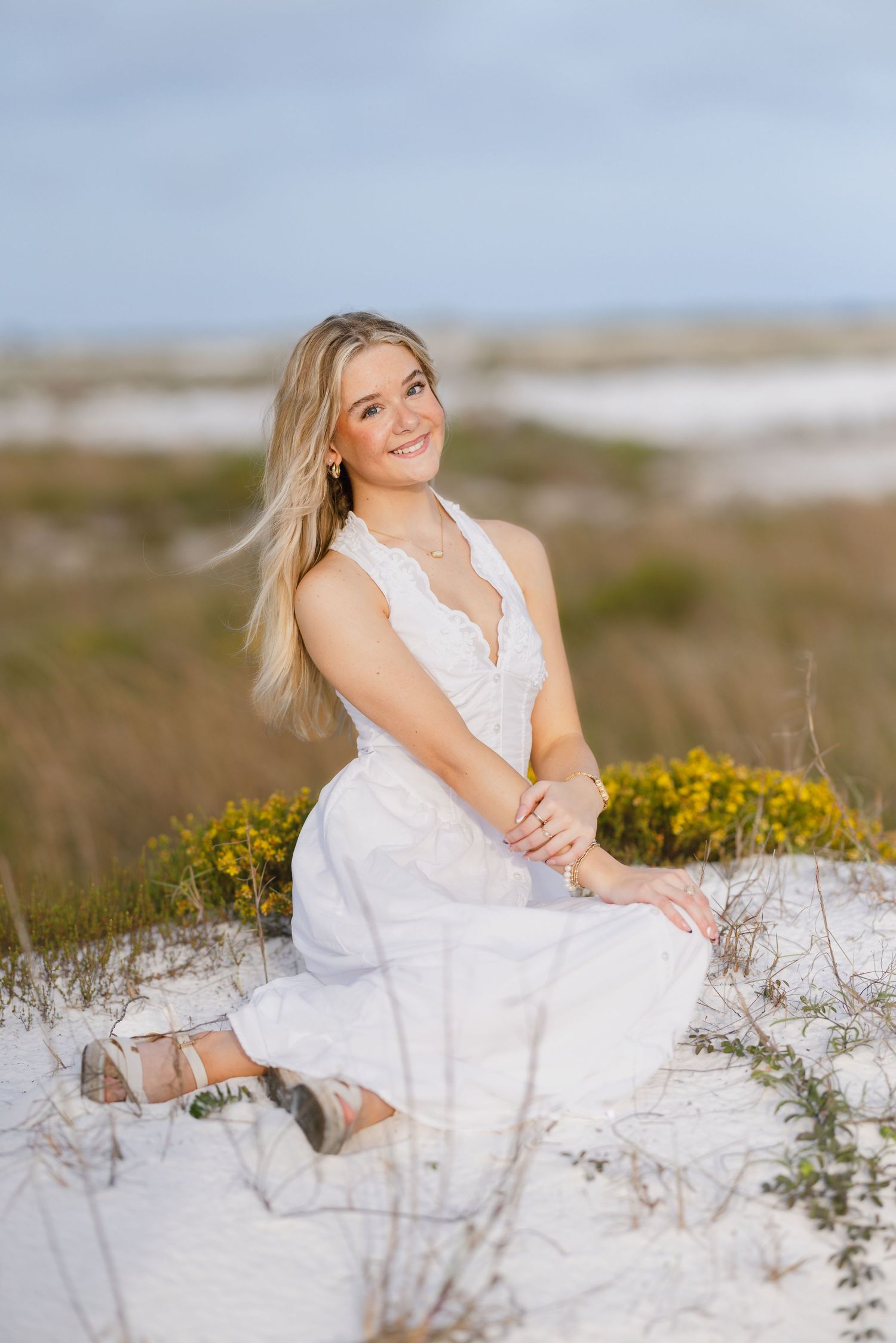 Smiling blonde woman in white dress sitting on sandy dunes with yellow wildflowers and coastal grasses in background.