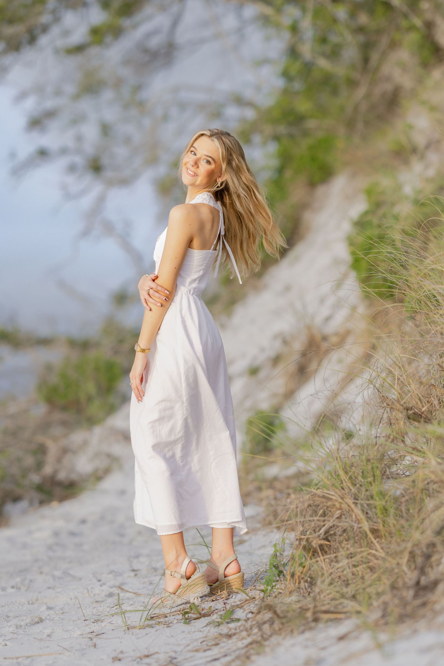 Woman in white sundress and wedge sandals poses on sandy path with coastal dunes and greenery in background.