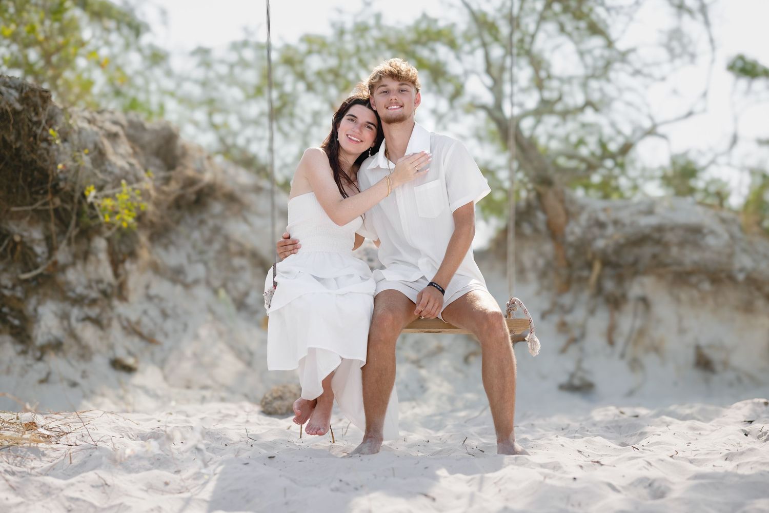 Happy couple sitting on a rope swing in sandy beach dunes with trees in the background.