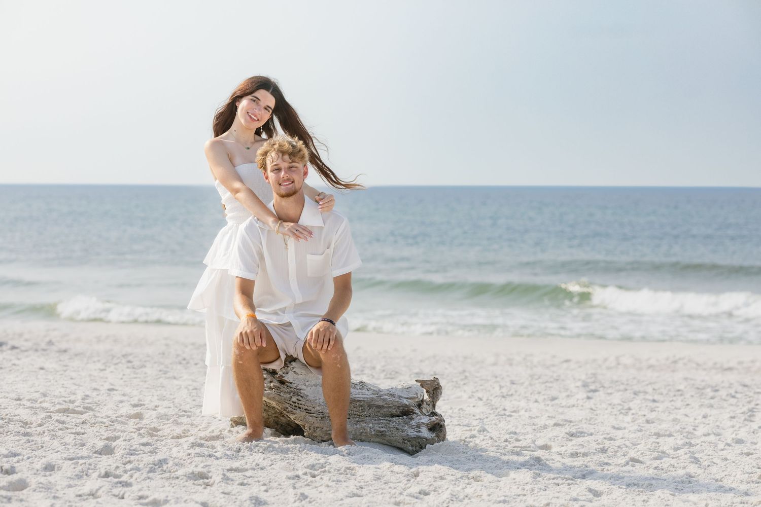 A young couple poses playfully on a driftwood log on a sunny white sand beach near the ocean.