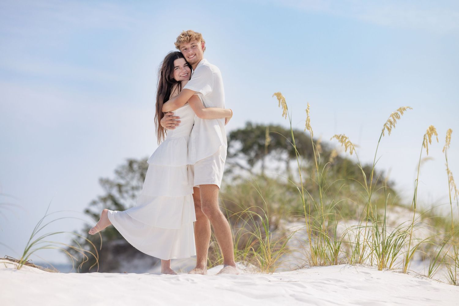 Couple in white outfits embracing on sandy beach dunes with sea oats and blue sky background.