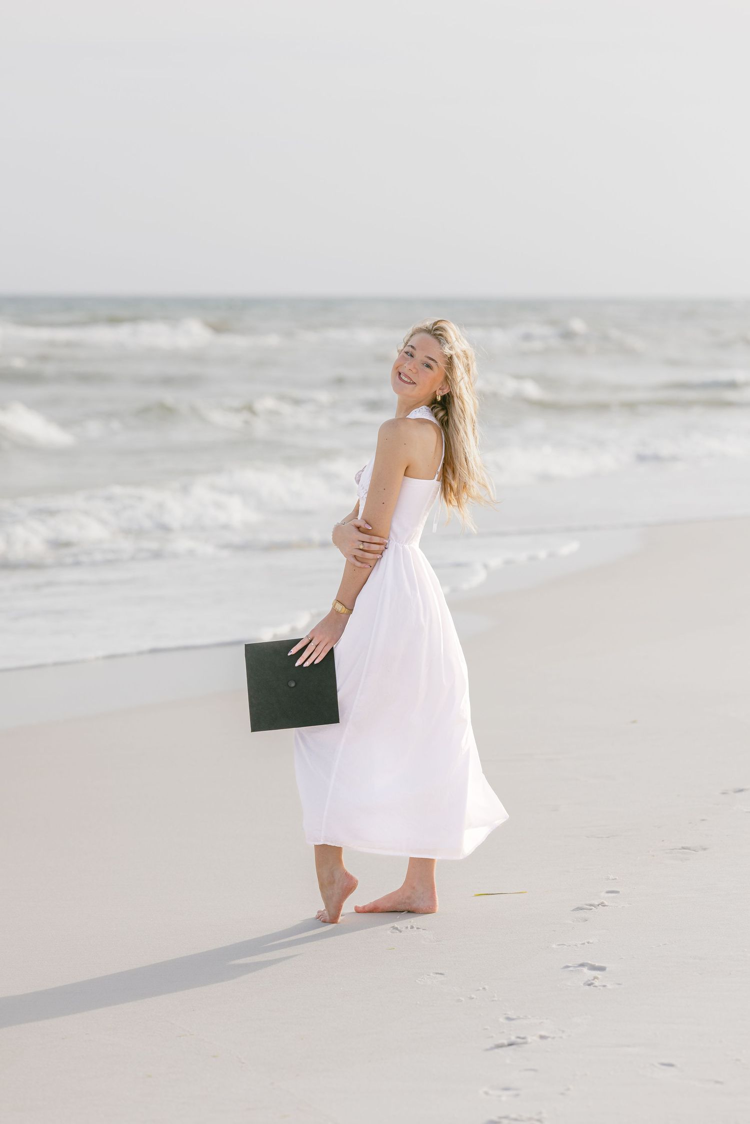 Smiling blonde woman in white off-shoulder dress holding diploma on sandy beach with ocean waves behind her.