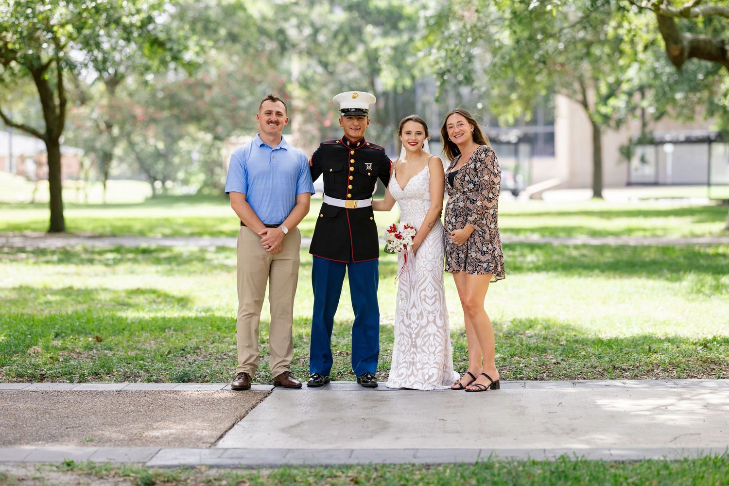 Marine in dress uniform poses with three family members outdoors on a sunny day near green trees and grass.