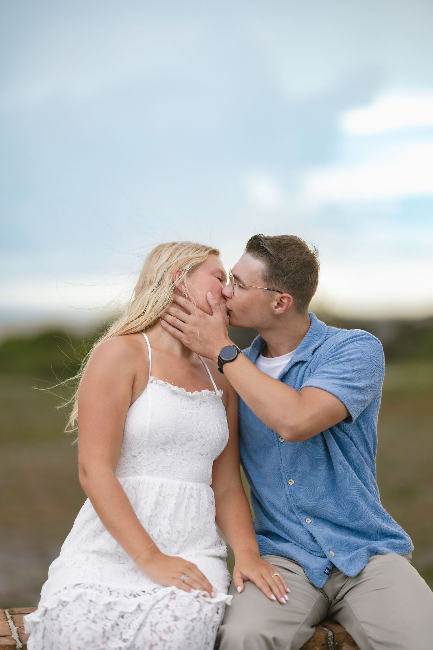 A couple shares a romantic kiss outdoors, woman in white lace dress and man in blue shirt, with a cloudy sky backdrop.