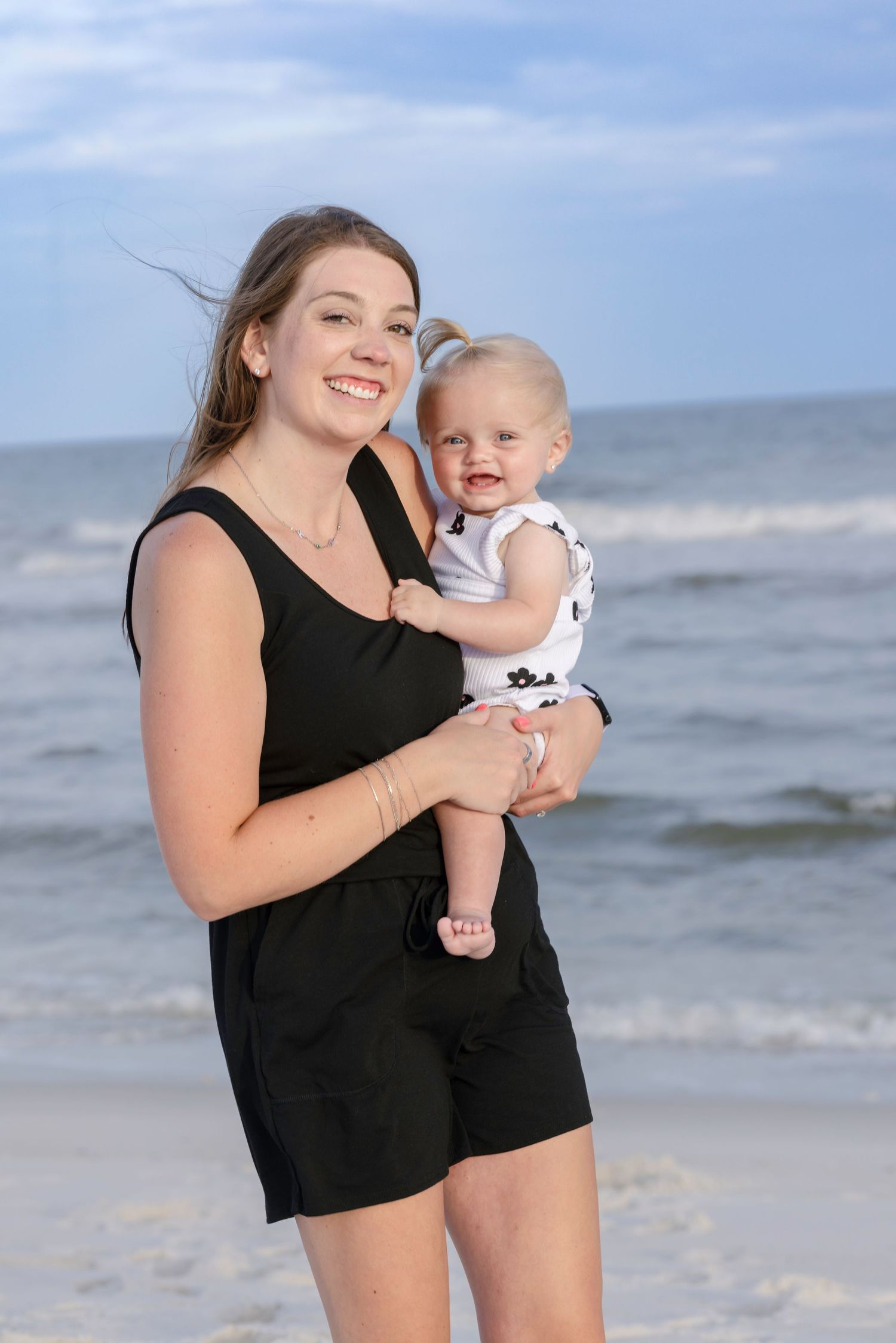 Smiling young woman in black romper holding a baby on a sunny beach with ocean waves in the background.