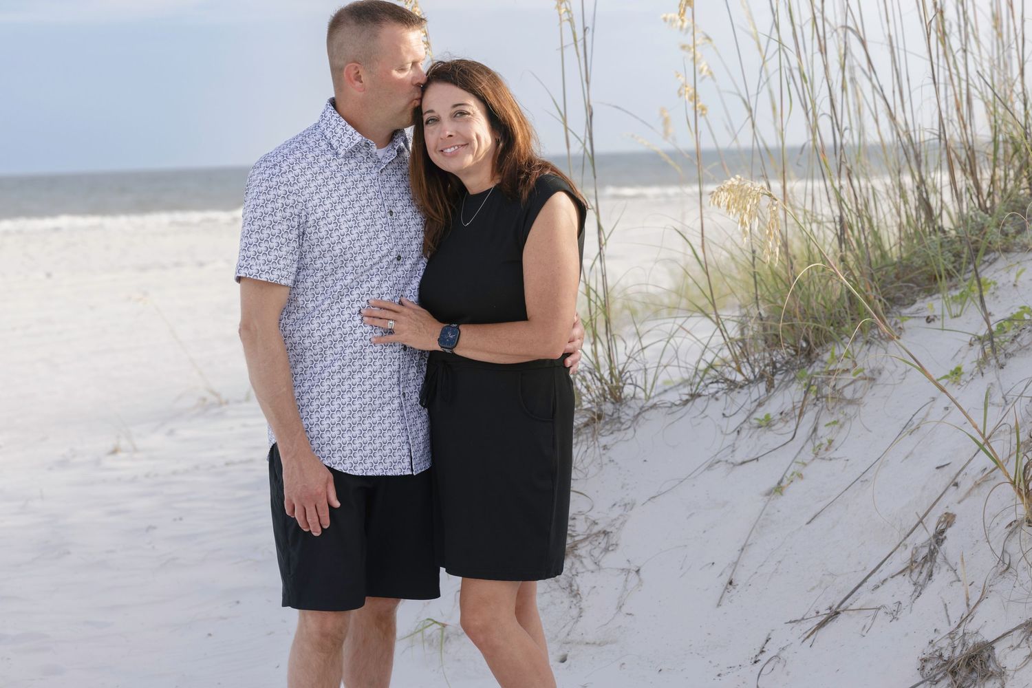 Couple embracing on a sandy beach with sea oats, man in plaid shirt and woman in black dress smiling.