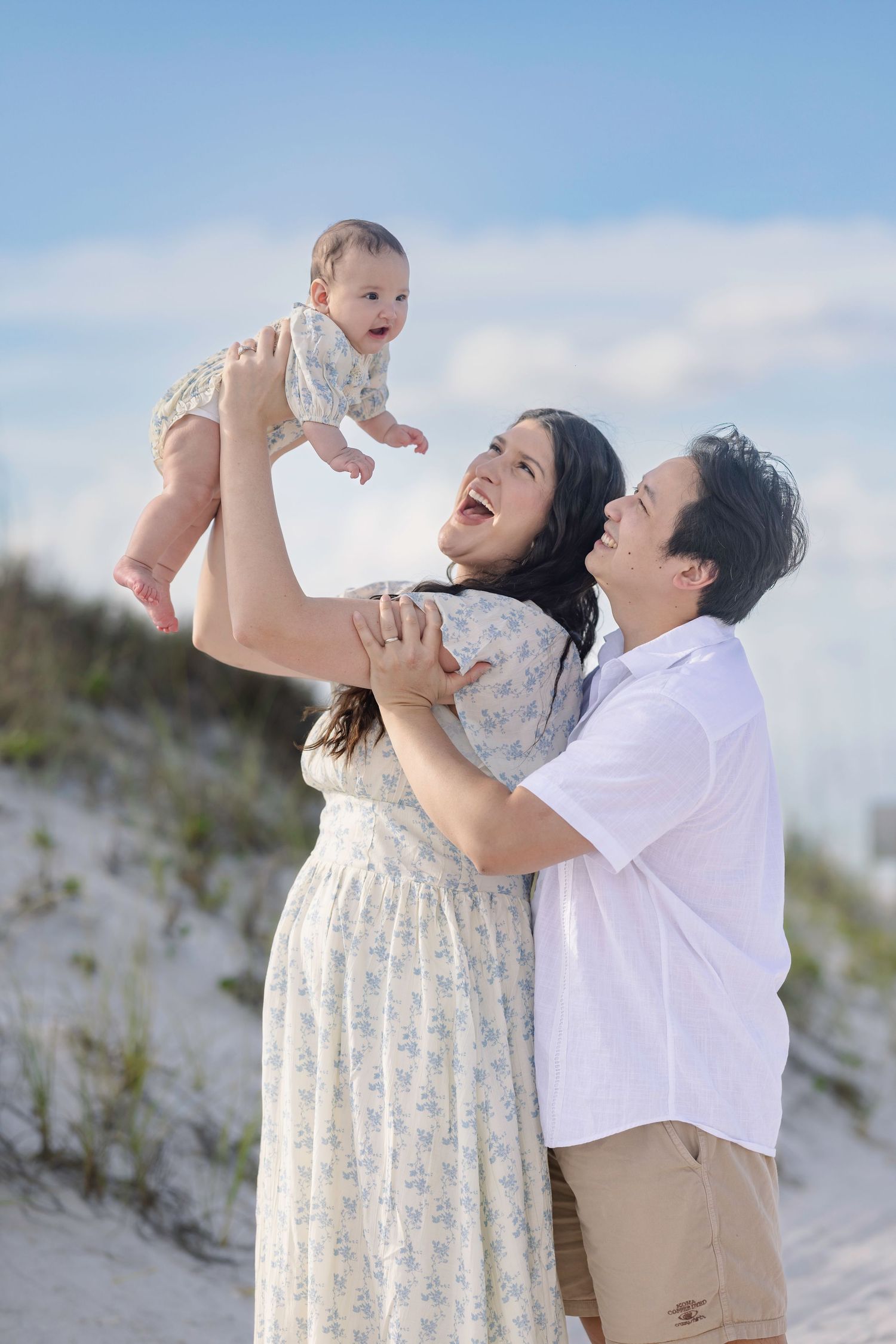Happy couple laughing on beach as mother lifts smiling baby in the air against blue sky.