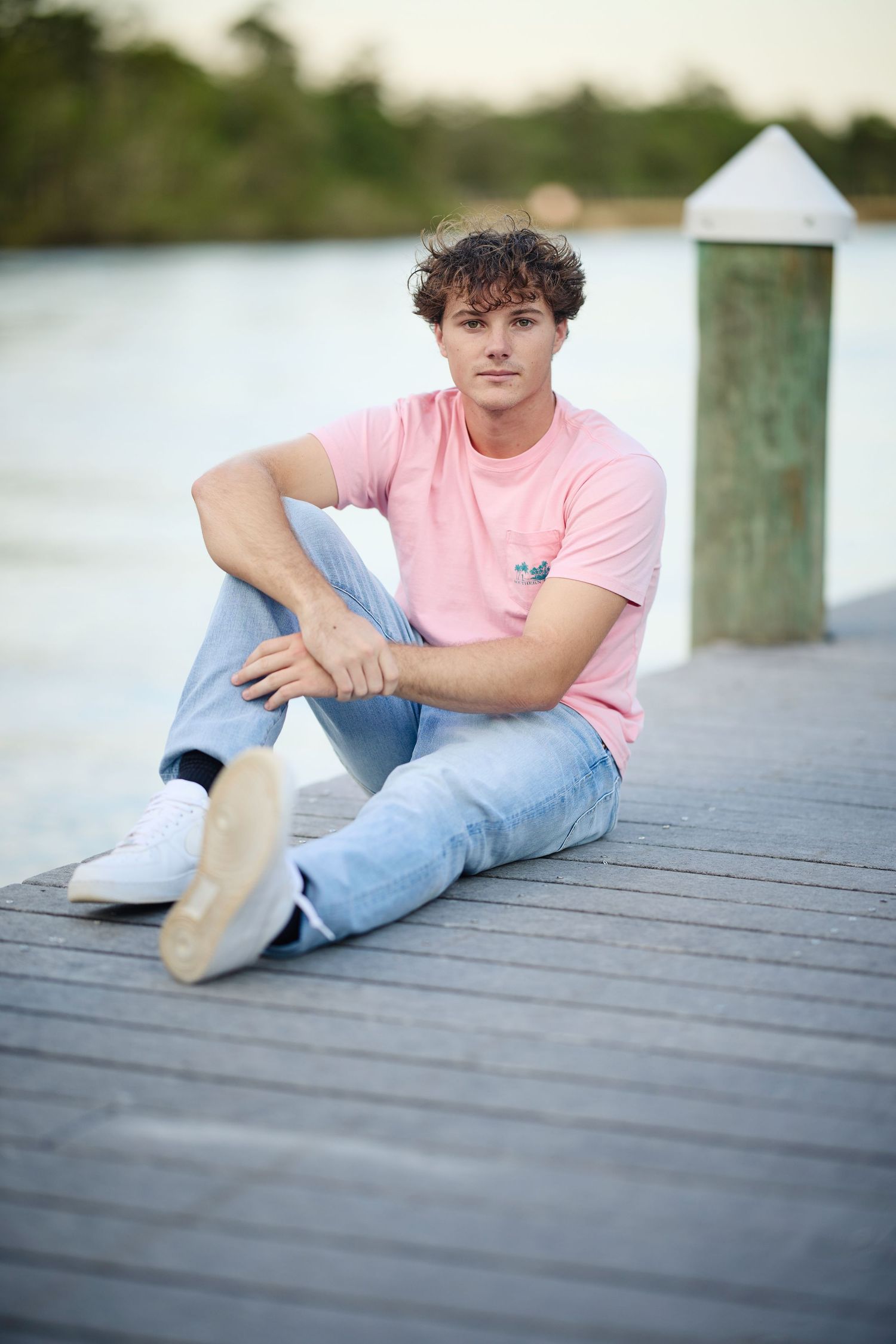 Casual portrait on wooden dock showing person in pink t-shirt and jeans sitting by waterfront with white pillar post.
