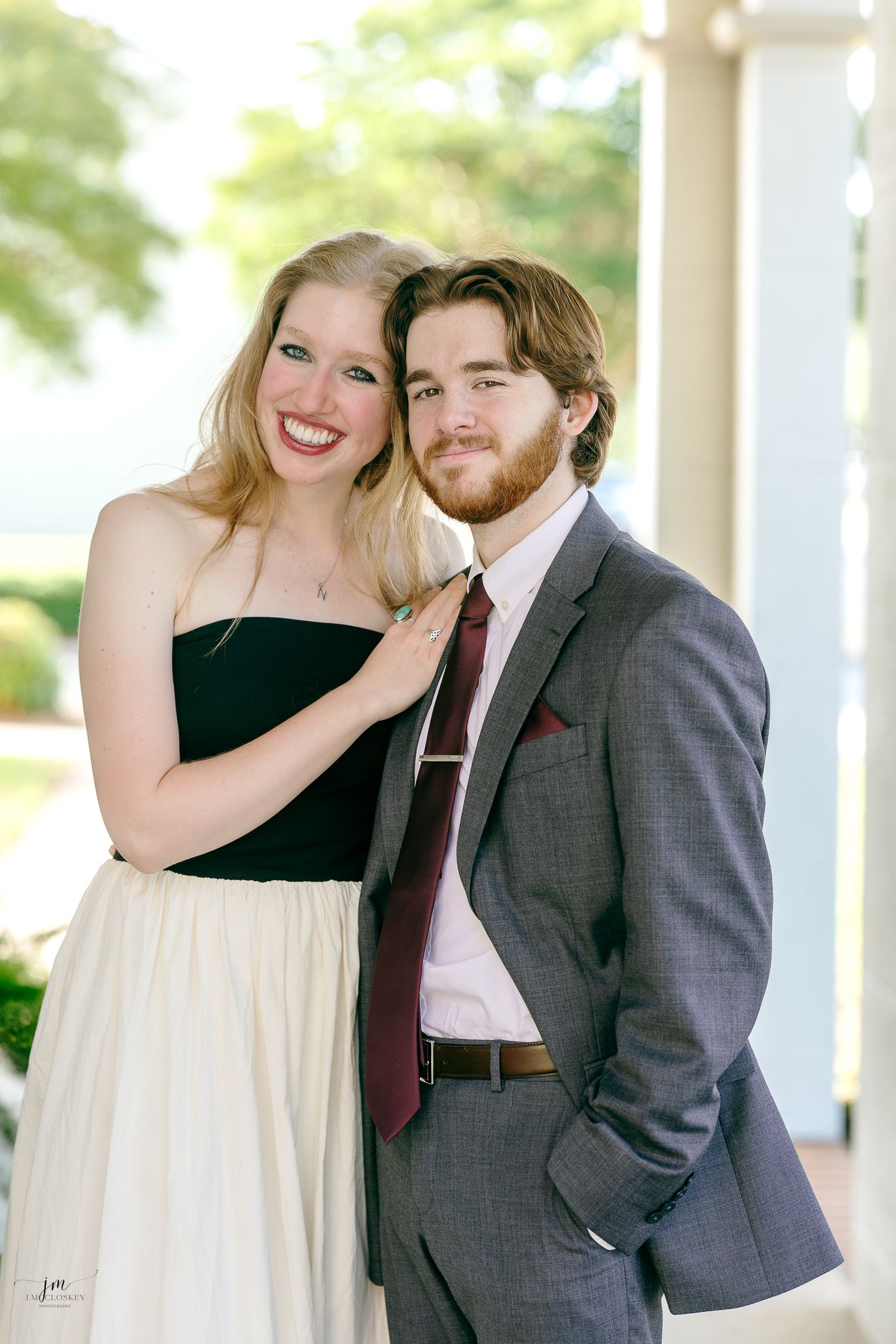 A couple poses together outdoors wearing formal attire - she in a black and cream dress, he in a grey suit.