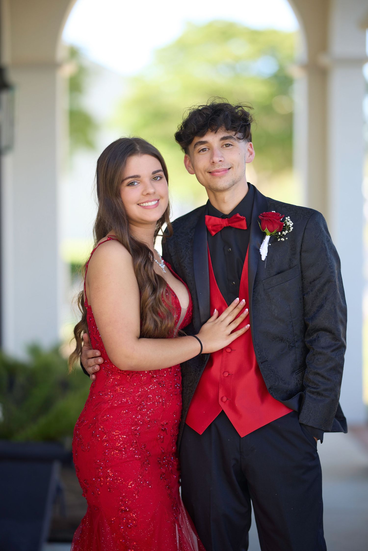 Couple in formal attire poses outdoors with person in red dress and black suit on porch setting.