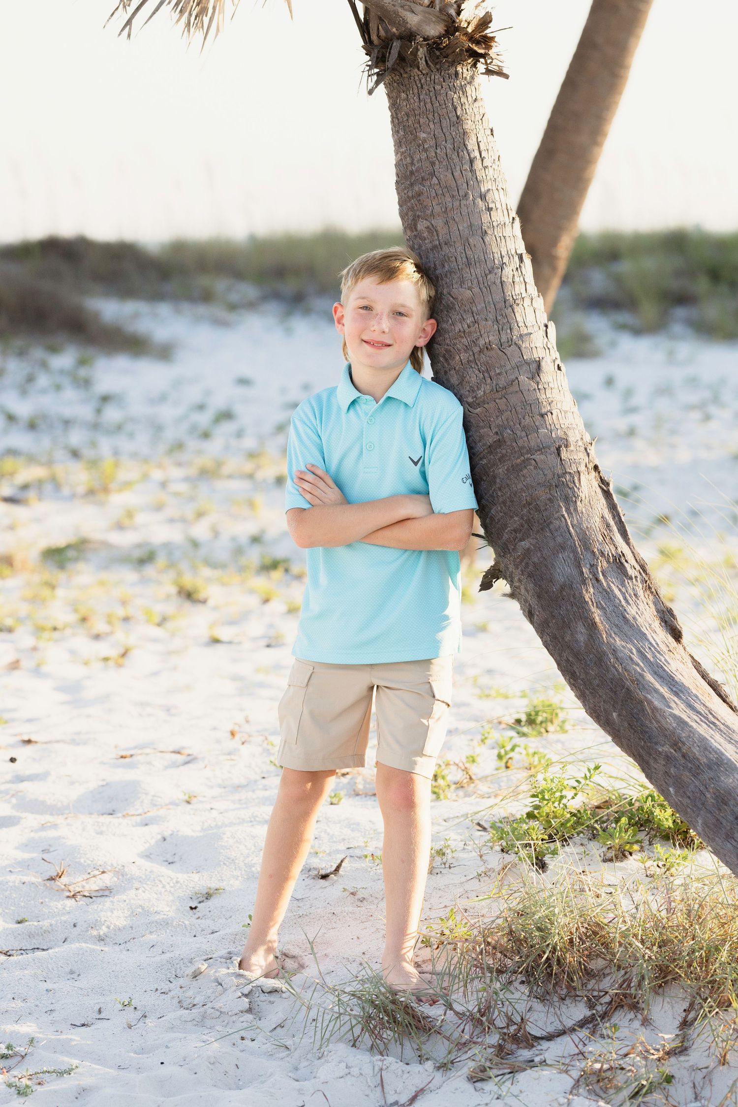 An aqua-colored shirt and khaki shorts create a beachy coastal vibe against white sand and a palm tree trunk.