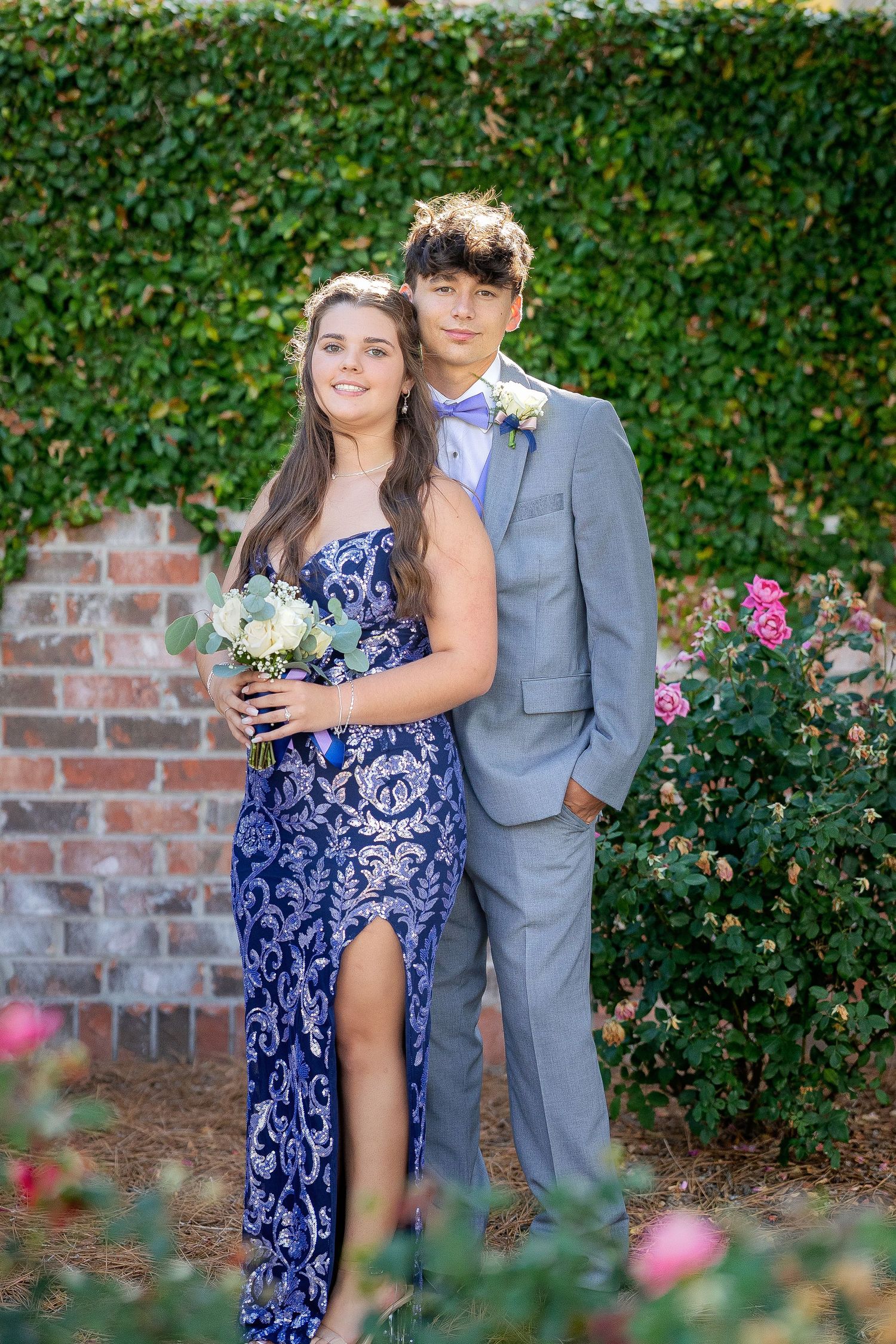 Couple in coordinating formal attire poses together against a lush green hedge backdrop with pink roses.