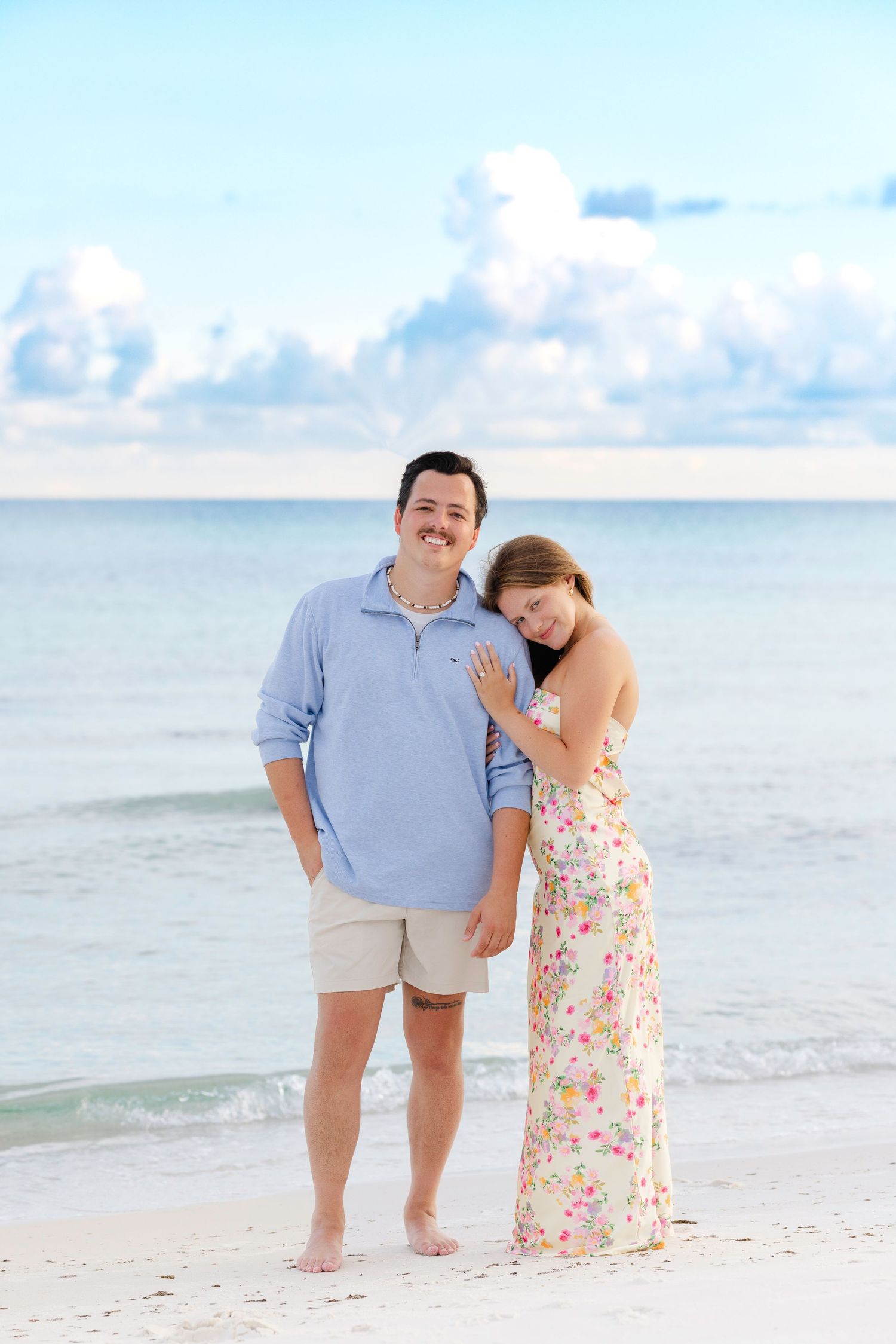 A romantic couple embraces while standing on a serene beach with cloudy blue skies in the background.