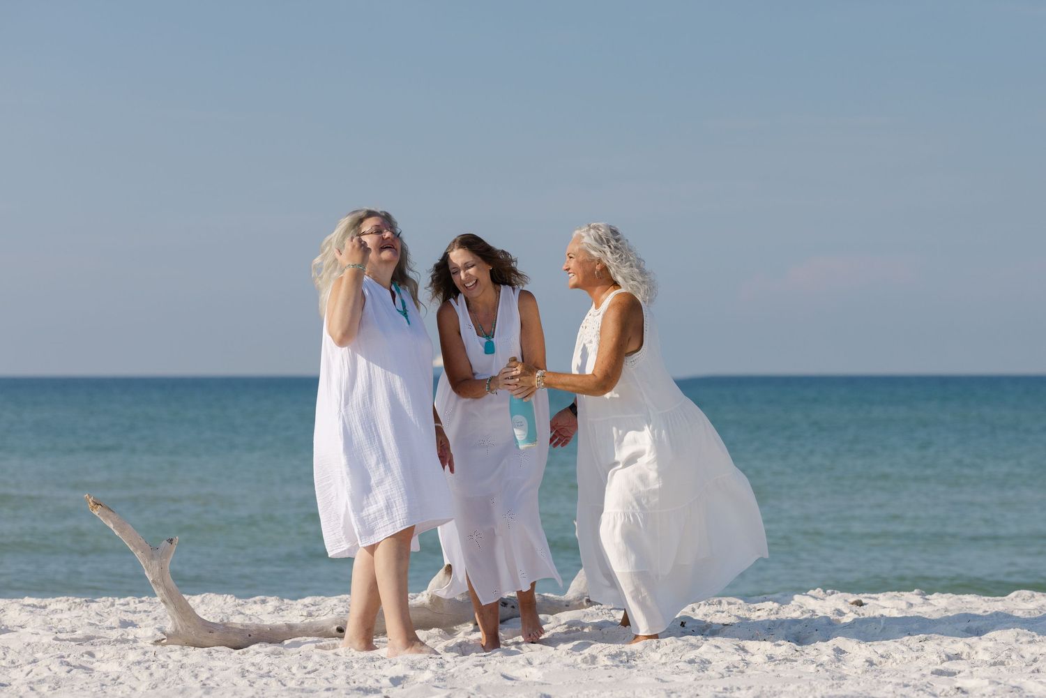 Three people in white dresses laugh together on a sunny beach with blue ocean waters in the background.