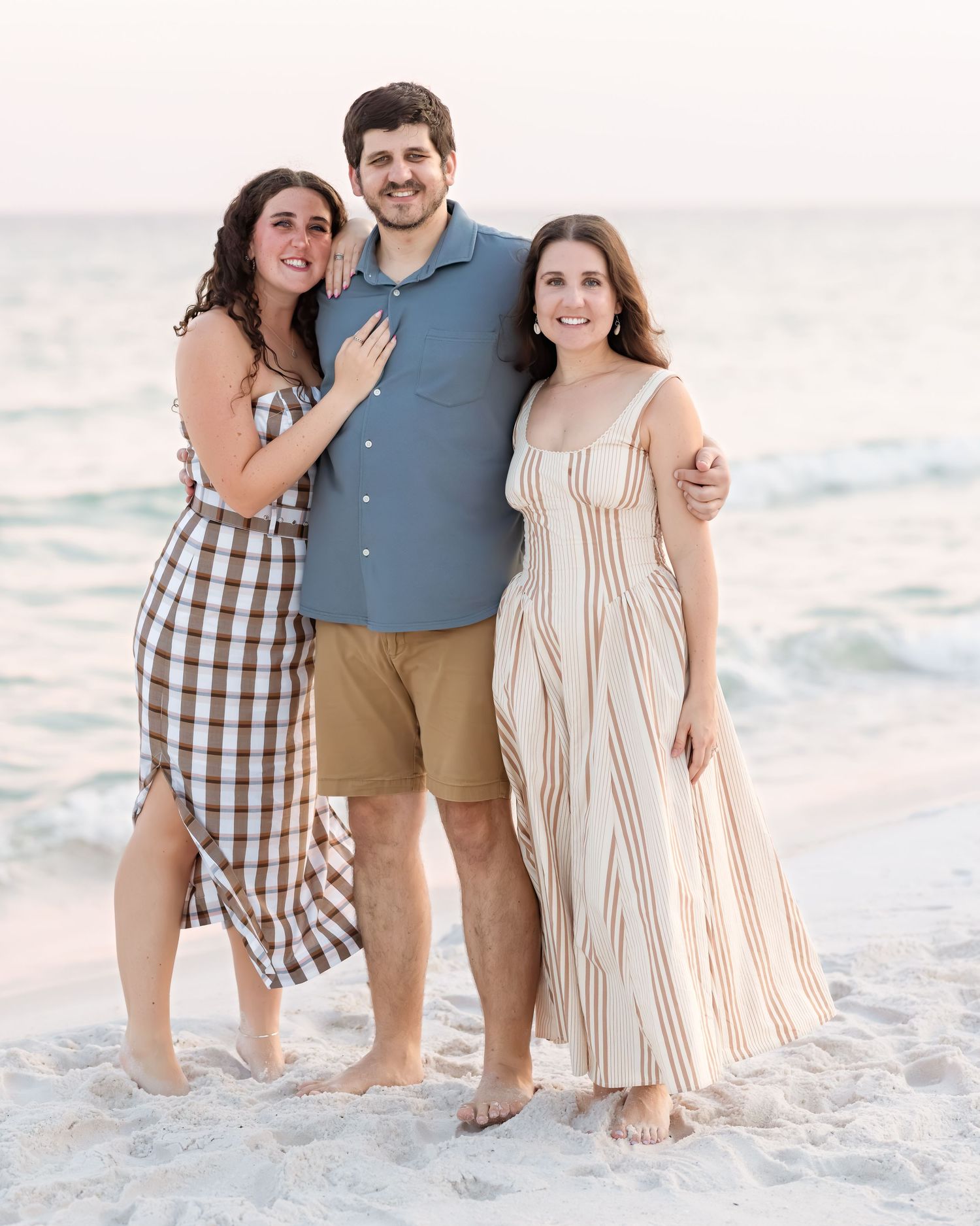 Three friends posing together on beach at sunset, woman in plaid dress, man in blue shirt, woman in striped maxi dress.