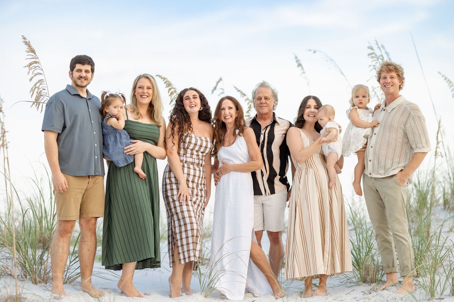 A joyful multigenerational family poses together on a sandy beach with sea oats in the background.