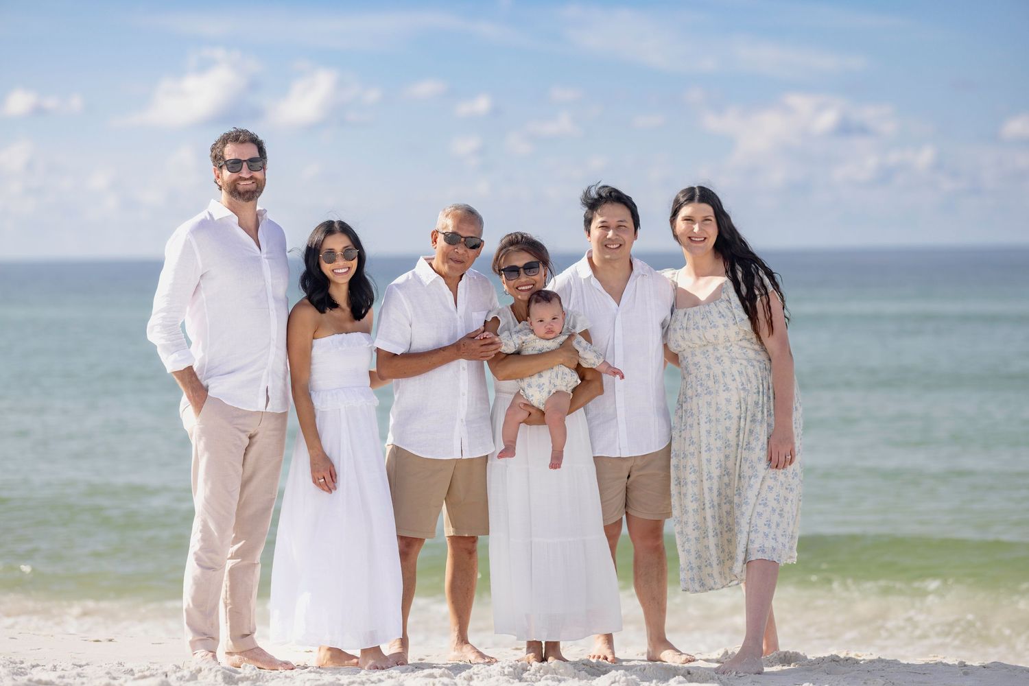 Family of seven posing together on a sunny beach with turquoise ocean waves in the background.