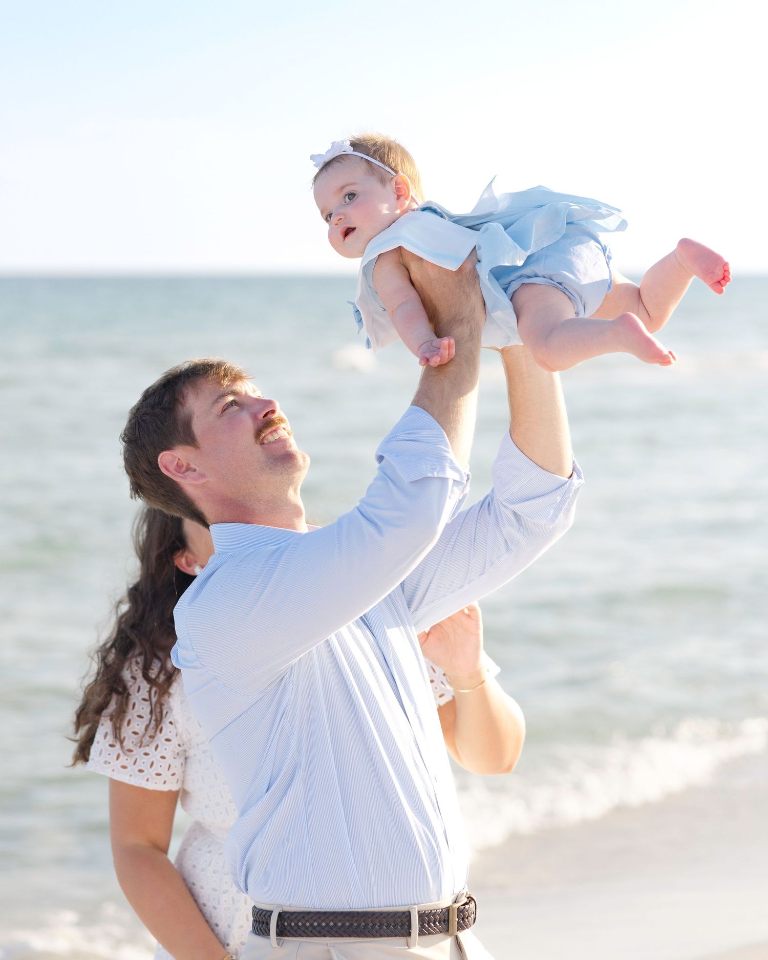 Family enjoying a playful moment at the beach with light blue dress and white outfits against ocean backdrop.