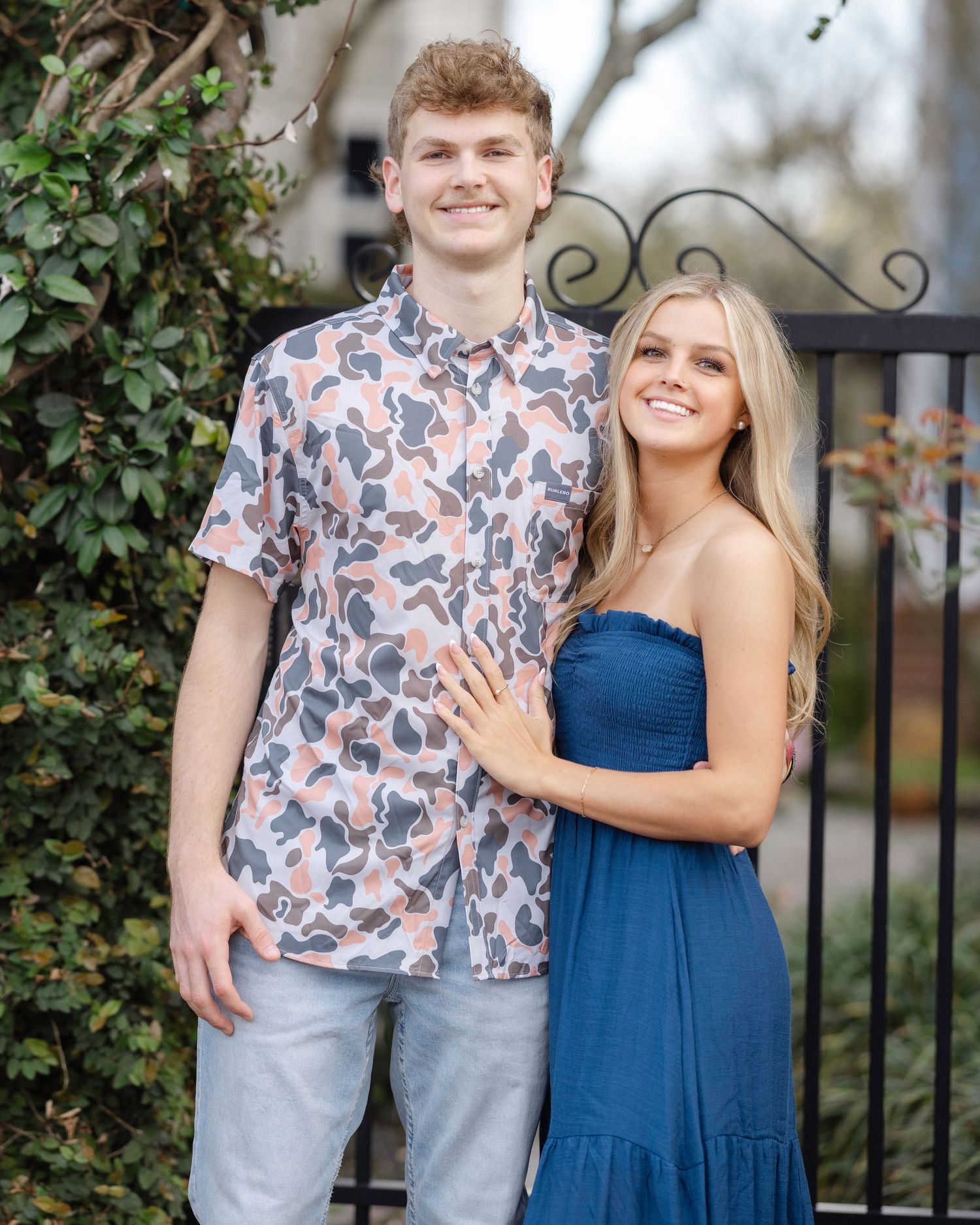 A couple poses together outdoors in coordinated outfits, with one wearing a floral shirt and the other in a navy dress.
