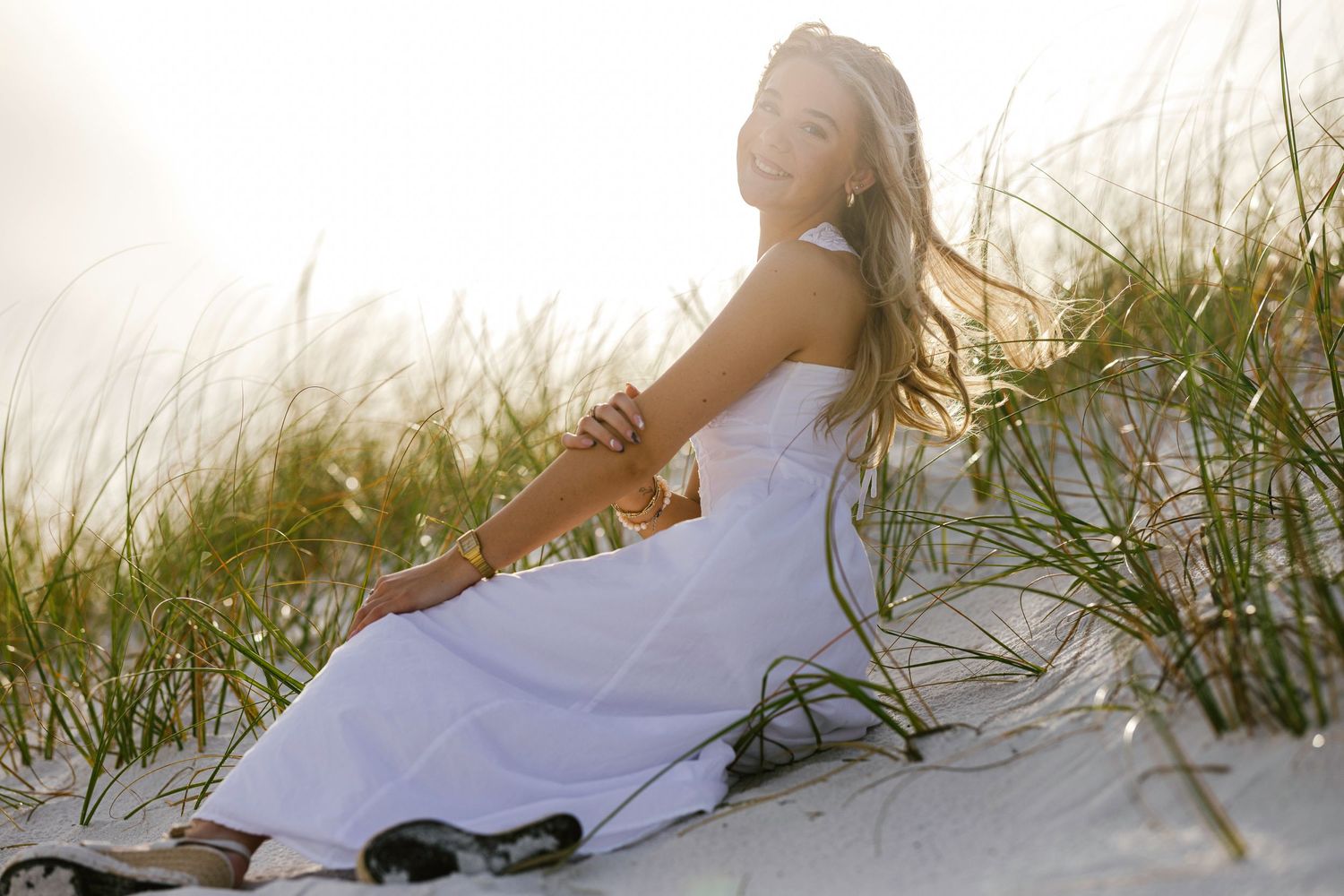 A woman in a white dress sits peacefully on a sandy beach dune at sunset among tall beach grass.