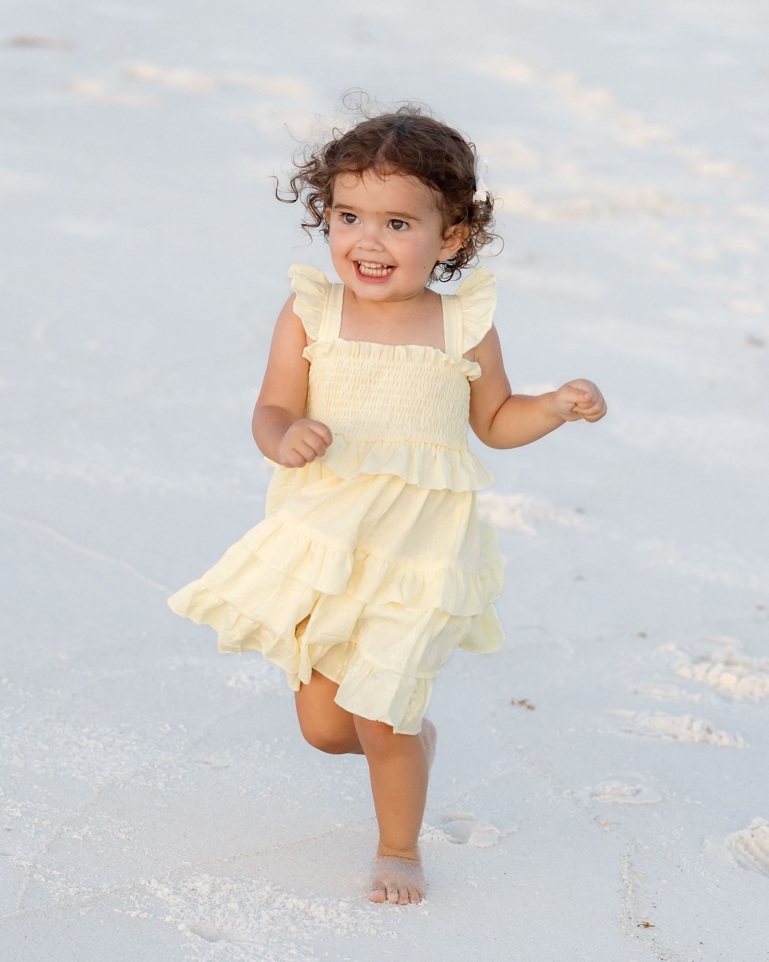 Happy toddler girl in yellow dress running barefoot on white sand beach.